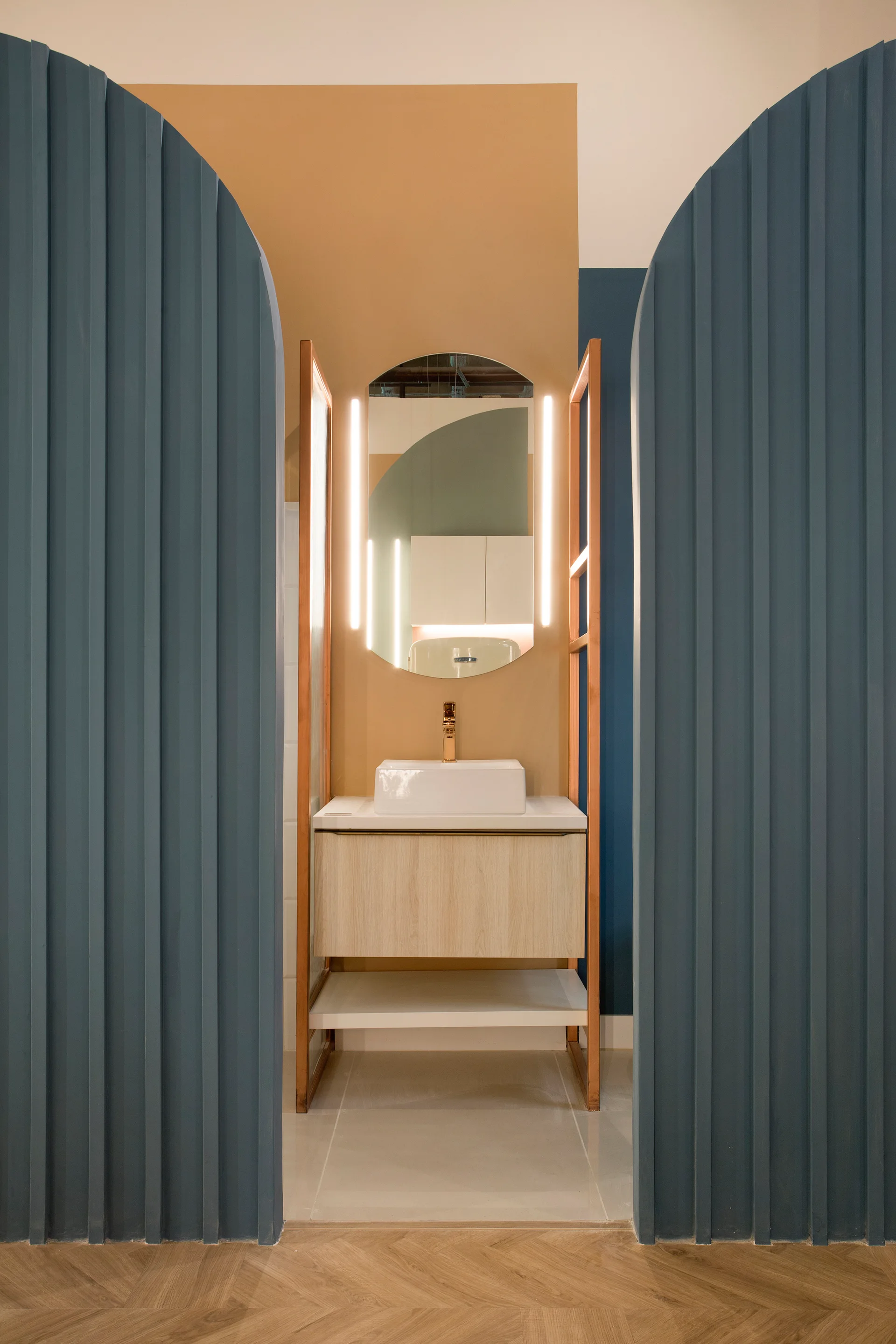 A light wood bathroom vanity with a white sink and backlit mirror is framed by two tall, arched blue fluted panels against a tan wall.