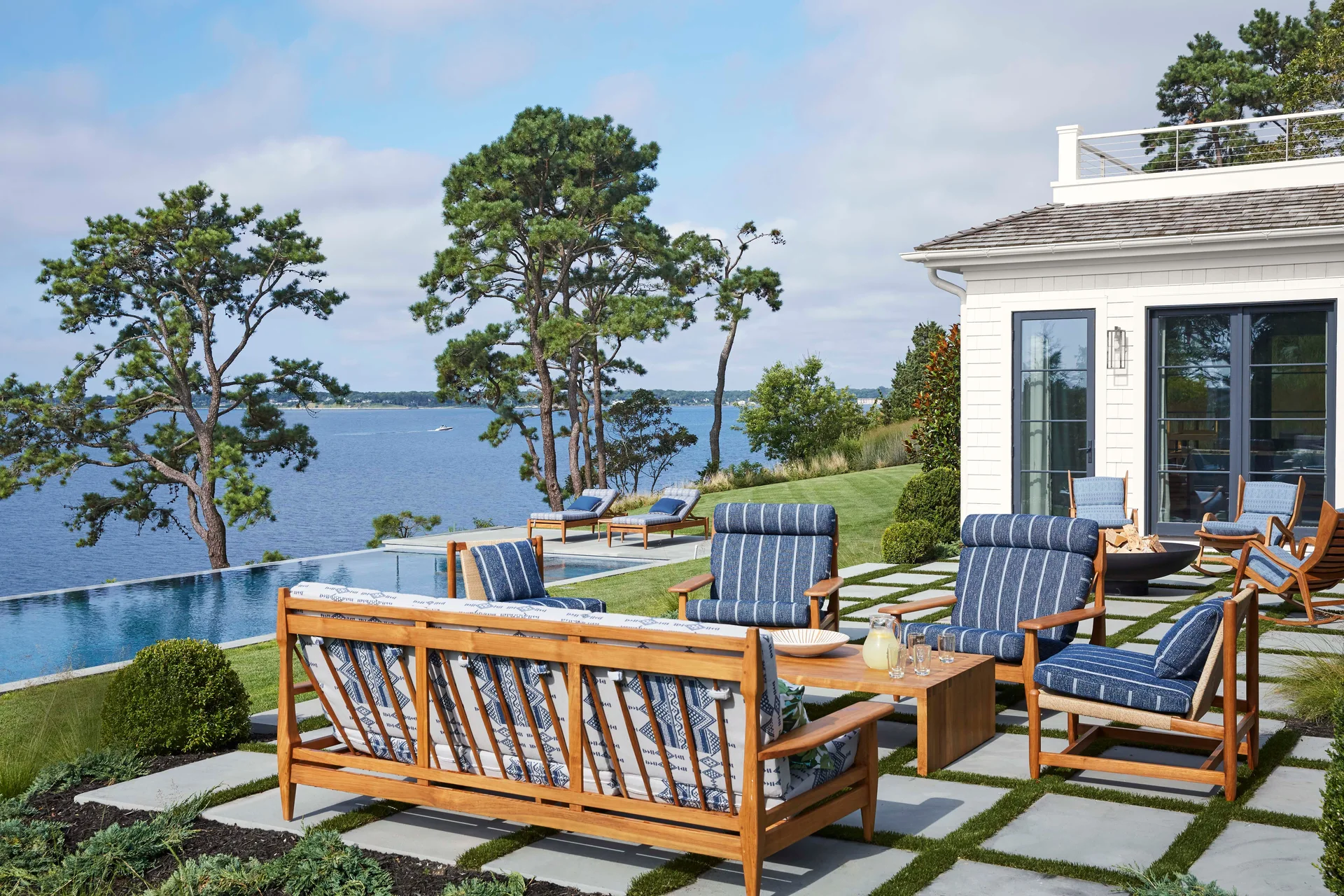 Blue and white patterned cushions top wooden patio furniture on gray stone pavers next to an infinity pool overlooking a body of water.