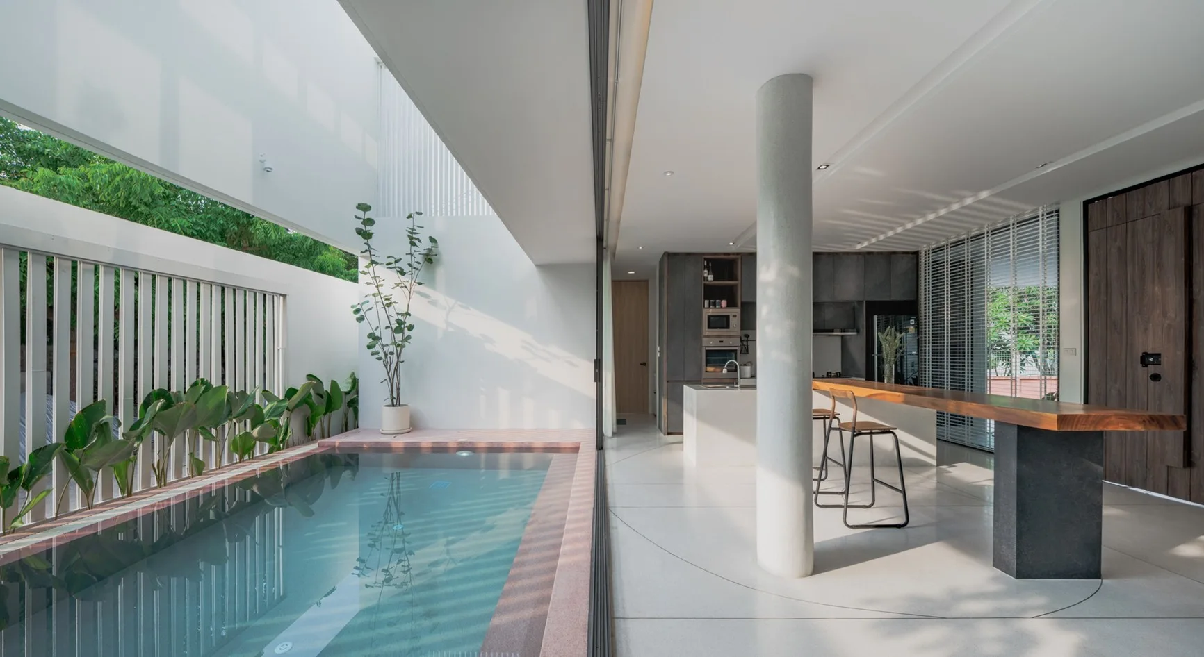 A minimalist modern kitchen with a long wood slab counter opens directly through a sliding glass wall to a narrow outdoor lap pool.