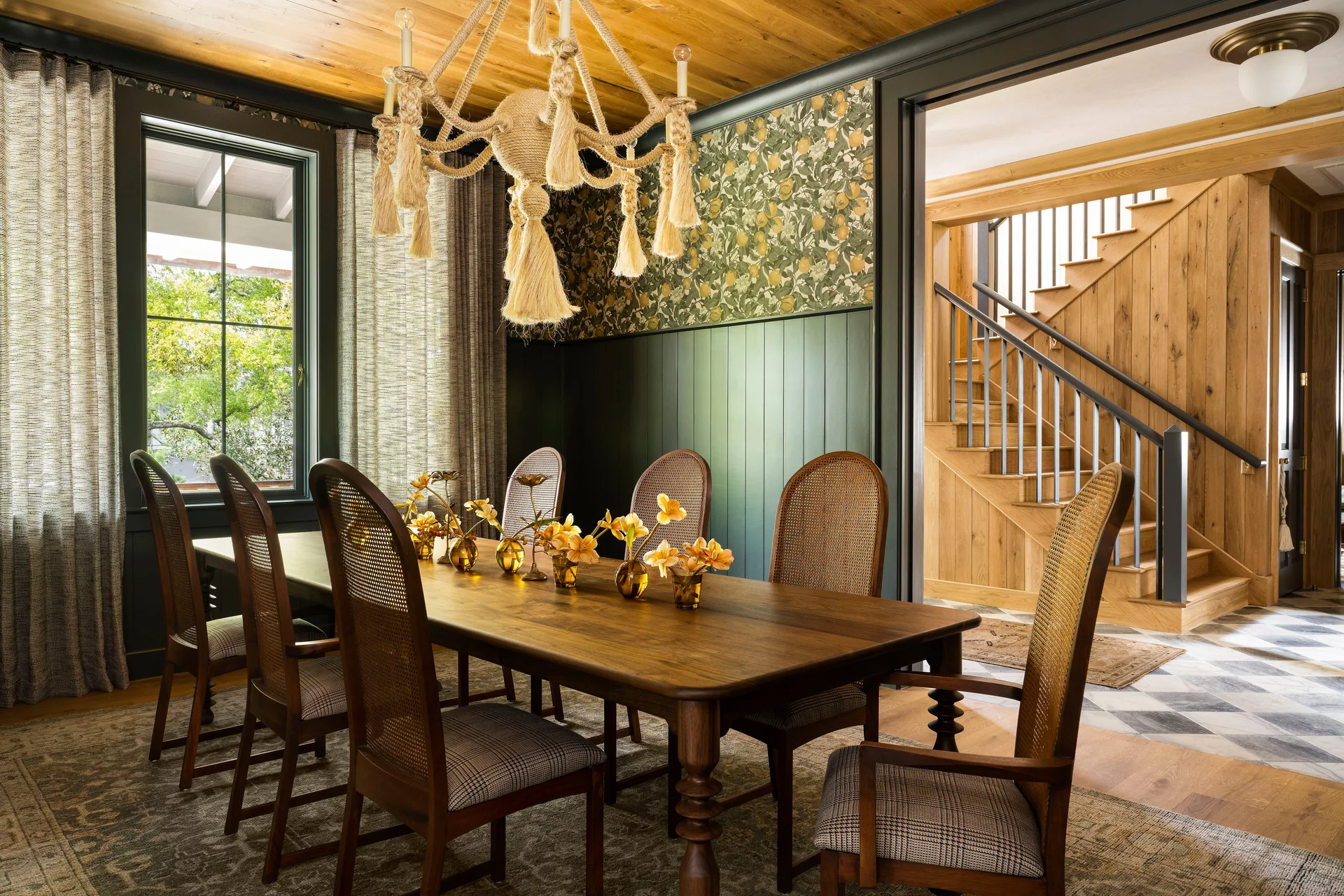 A wooden dining table and cane-backed chairs sit beneath a rope chandelier in a room with dark green paneled walls and patterned wallpaper.