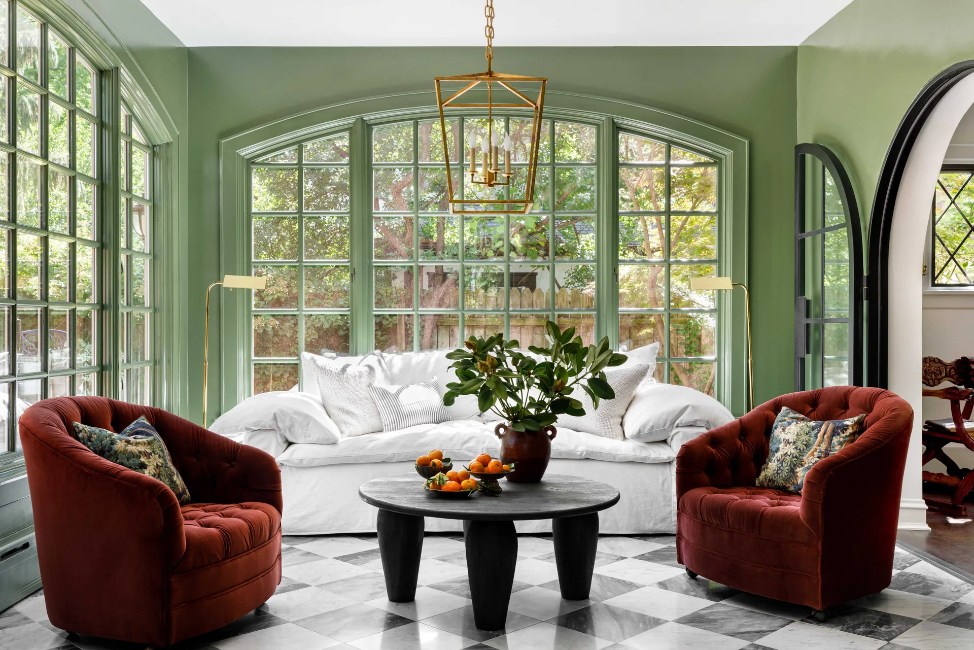 A sunlit room with sage green walls, a white sofa, two rust-red tufted velvet armchairs, and a black and white checkered marble tile floor.
