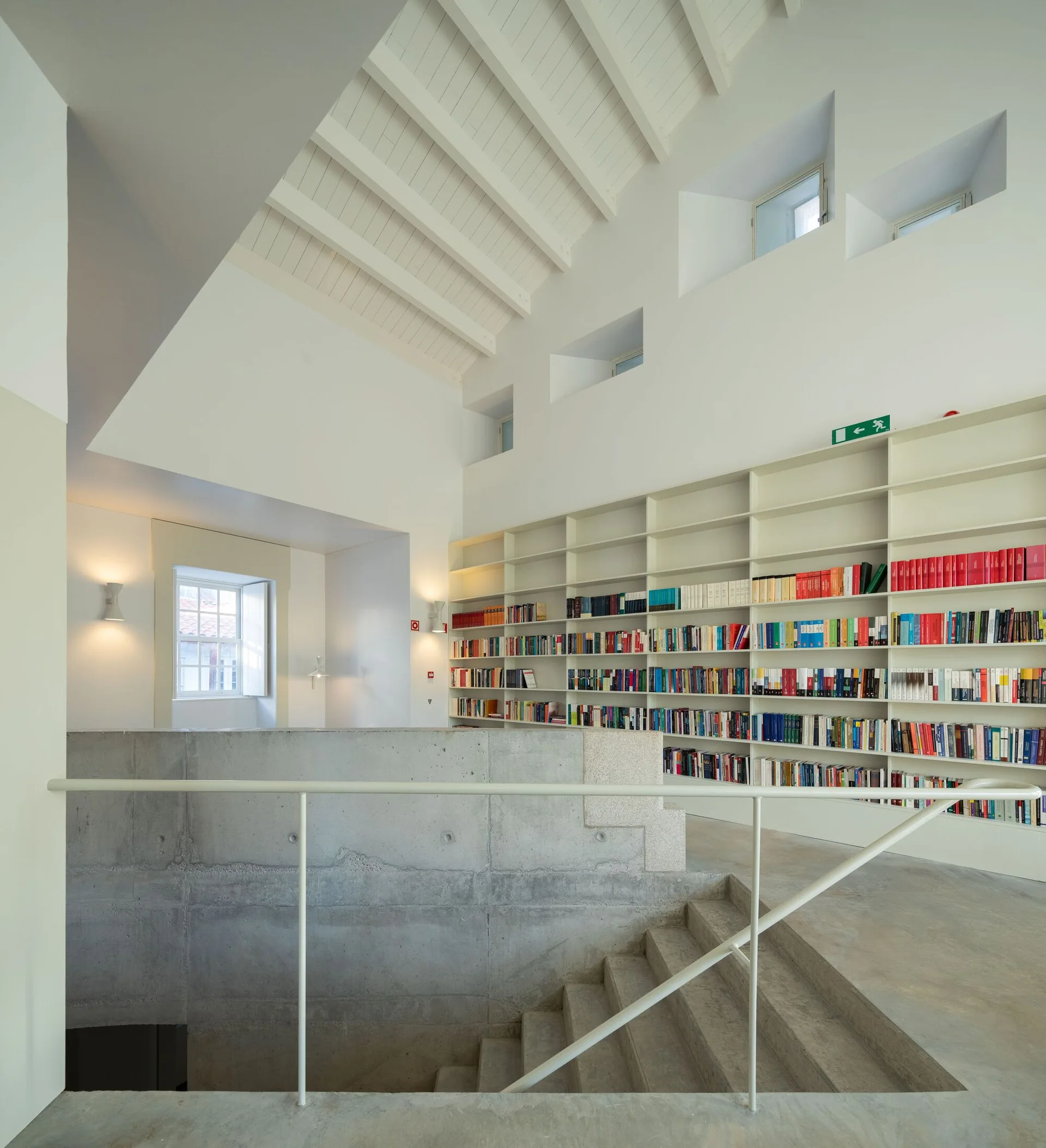 A modern library space featuring a massive white built-in bookshelf, concrete stairs with a white metal railing, and an exposed rafter ceiling.
