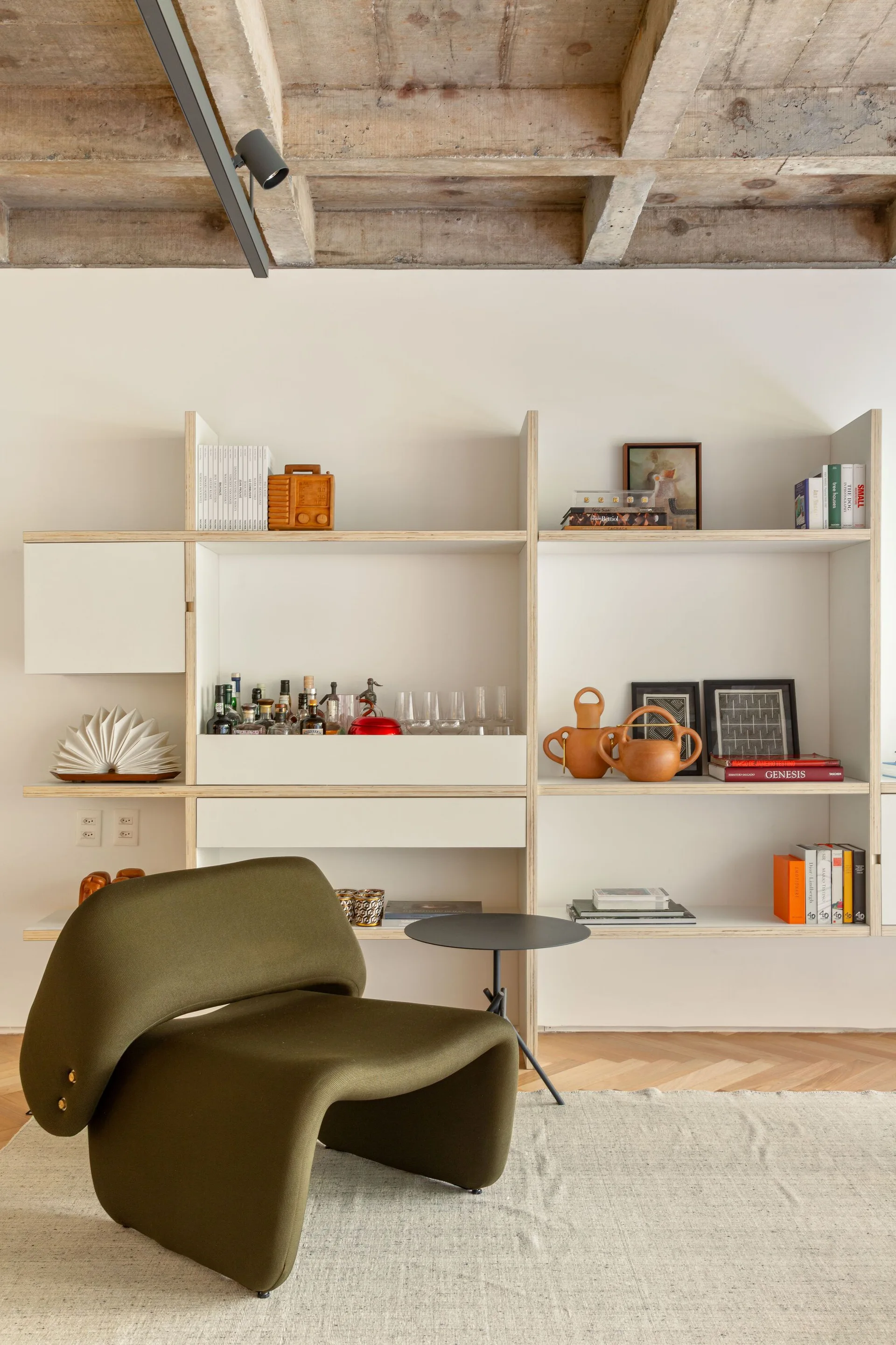 An olive green sculptural armchair sits on a light rug in front of a custom plywood shelving unit under an exposed concrete ceiling.