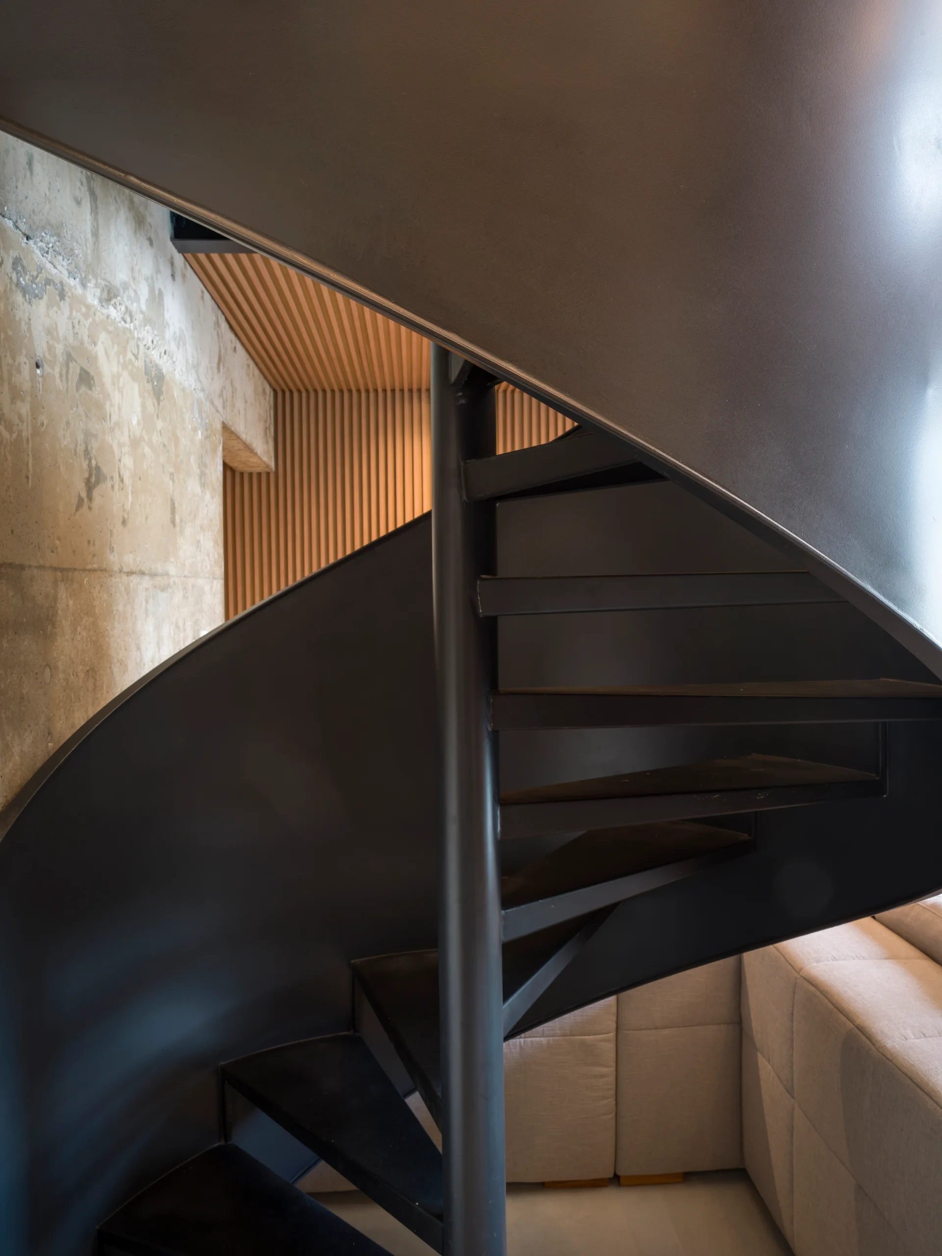 A black metal spiral staircase stands in front of a raw concrete wall and a textured vertical wood slat wall near a light beige sofa.