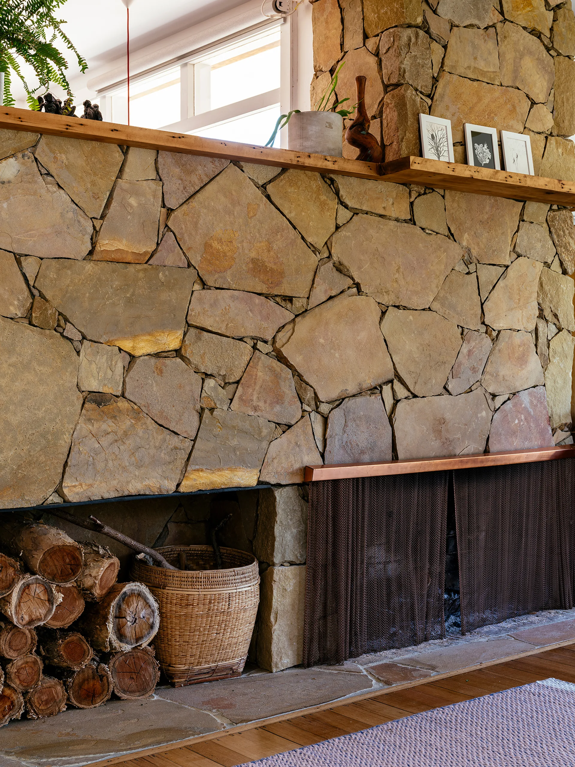 A large fireplace featuring irregular tan stone cladding, a natural wood mantel shelf, a woven log basket, and a dark mesh metal screen.