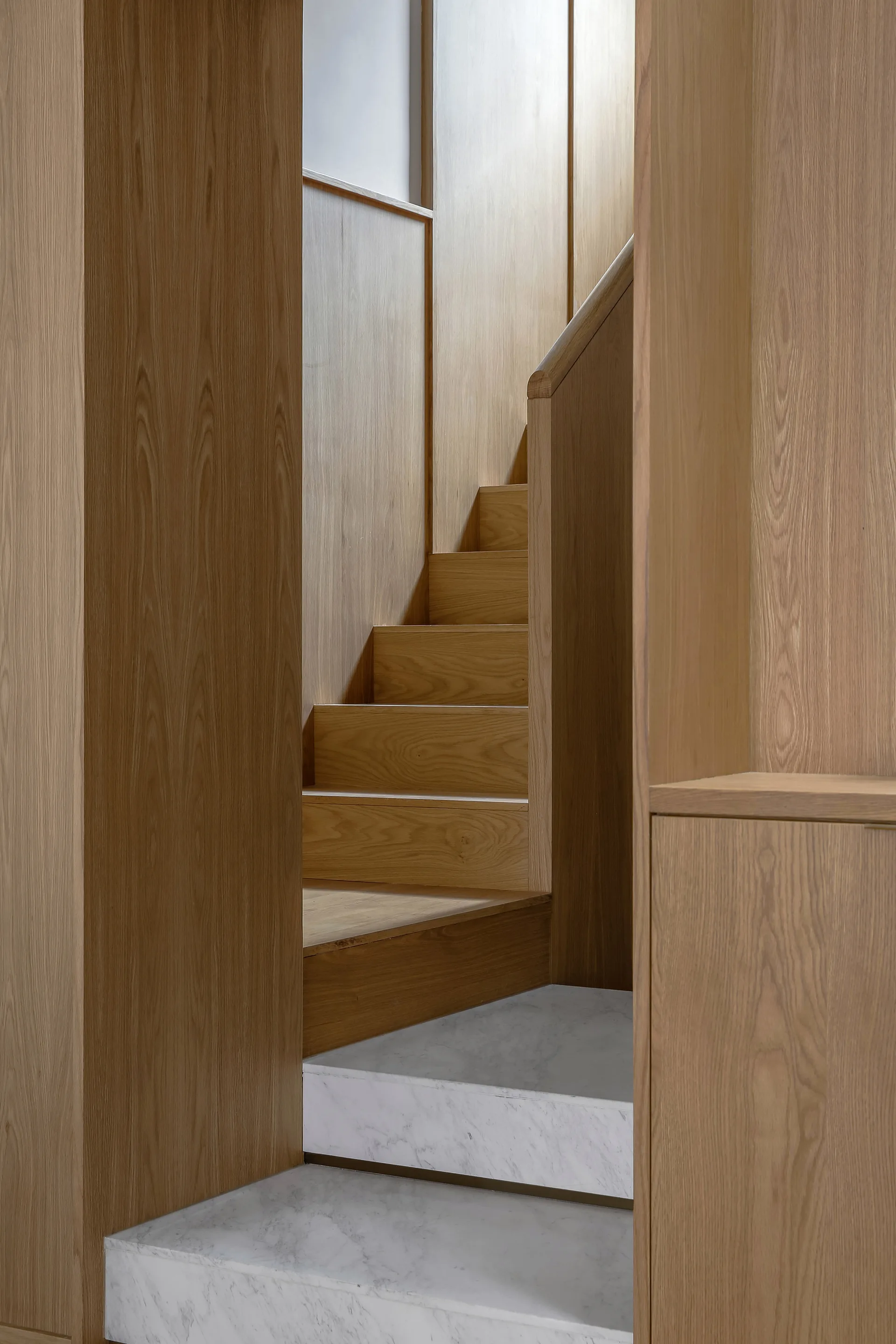 A minimalist interior staircase features warm wood wall paneling and matching steps rising from a thick, white veined marble base.