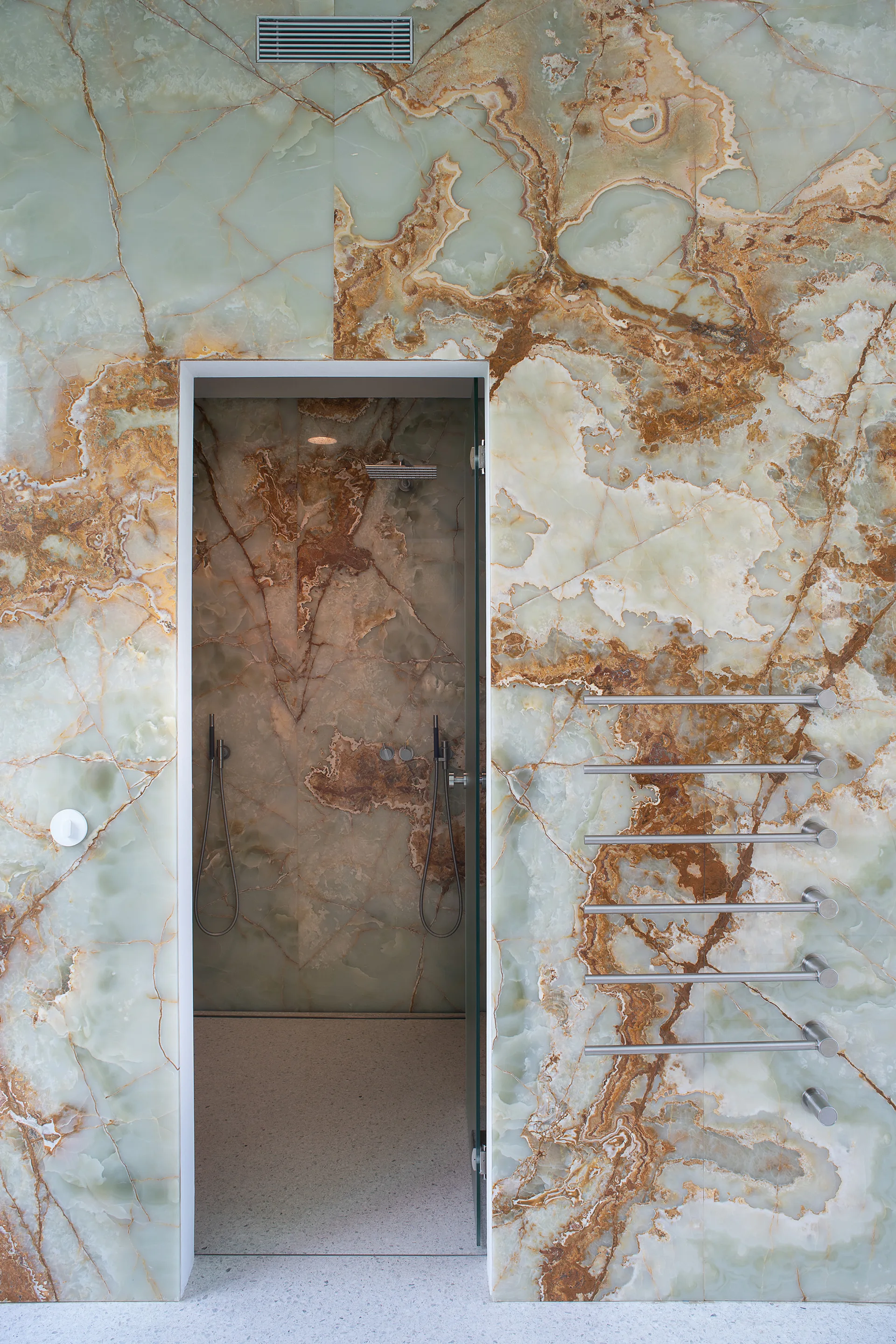 A modern bathroom features polished green onyx walls with amber veining, a glass-door shower, and built-in metal towel warming bars.