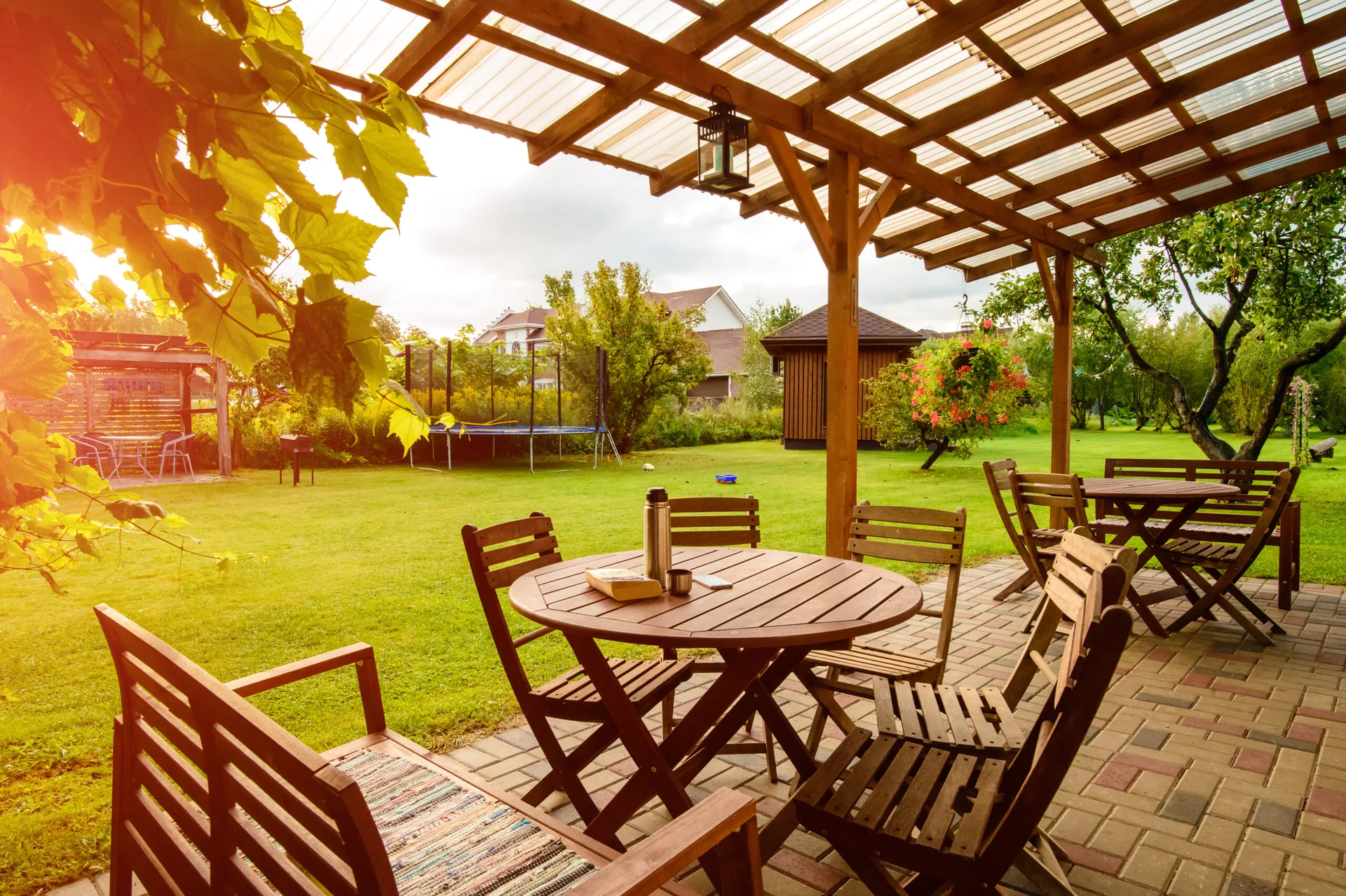 A covered stone paver patio features a wooden dining table and chairs under a timber pergola overlooking a grassy backyard lawn.