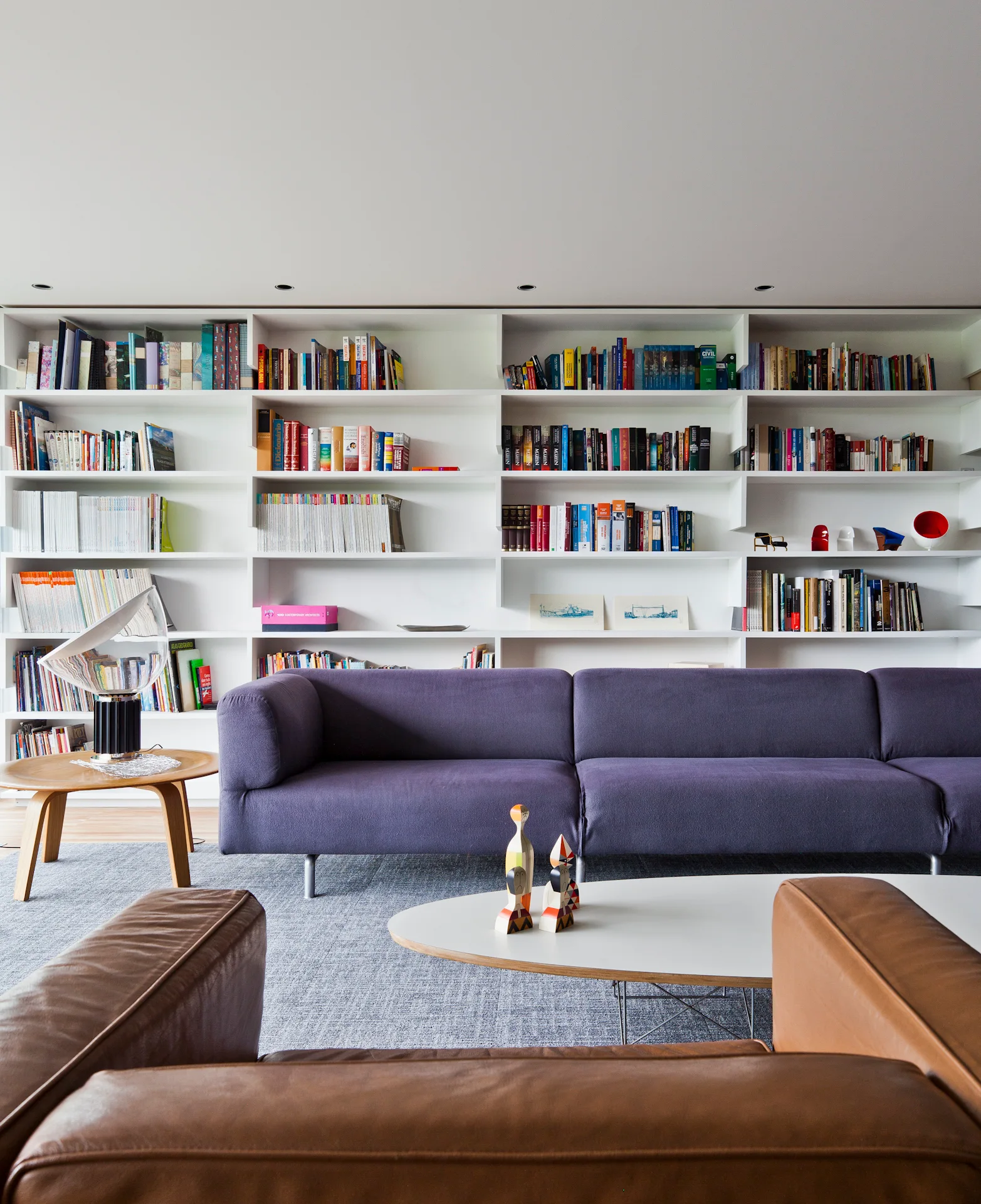A modern living room features a purple sofa placed in front of a white wall-to-wall built-in bookshelf filled with colorful books.