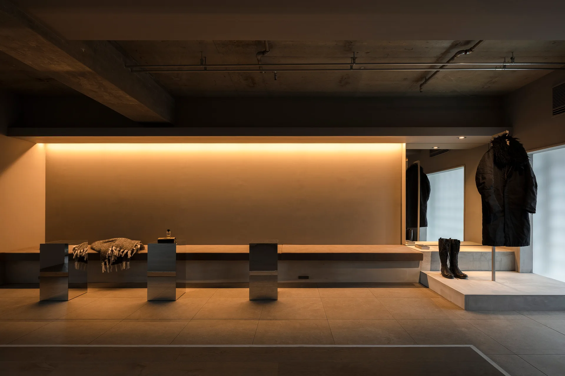 A minimalist industrial room features a long built-in upholstered bench, mirrored cube tables, concrete flooring, and warm, hidden cove lighting.