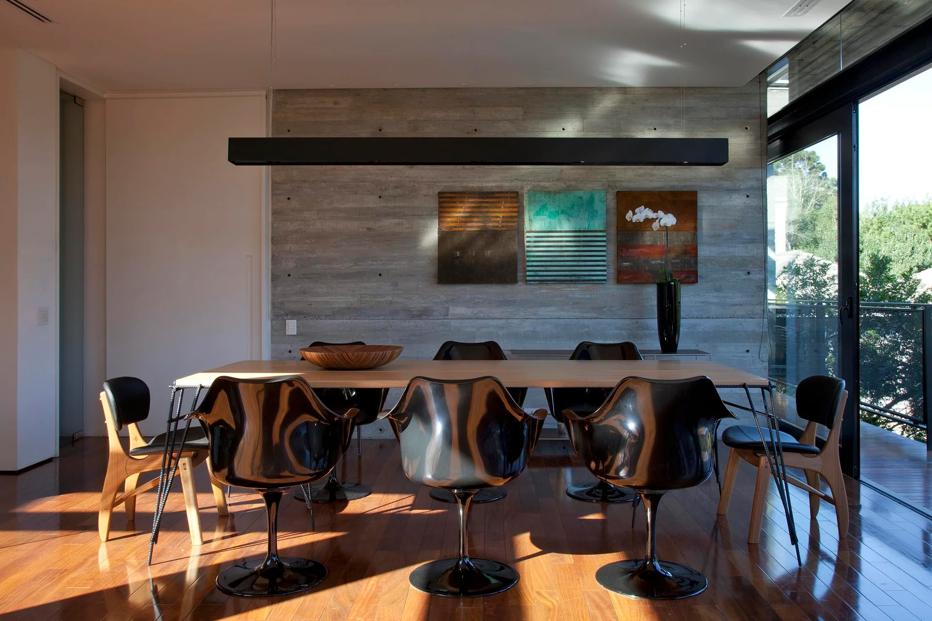 A modern dining room featuring a light wood table with black sculptural chairs against a board-formed concrete wall with three colorful art panels.