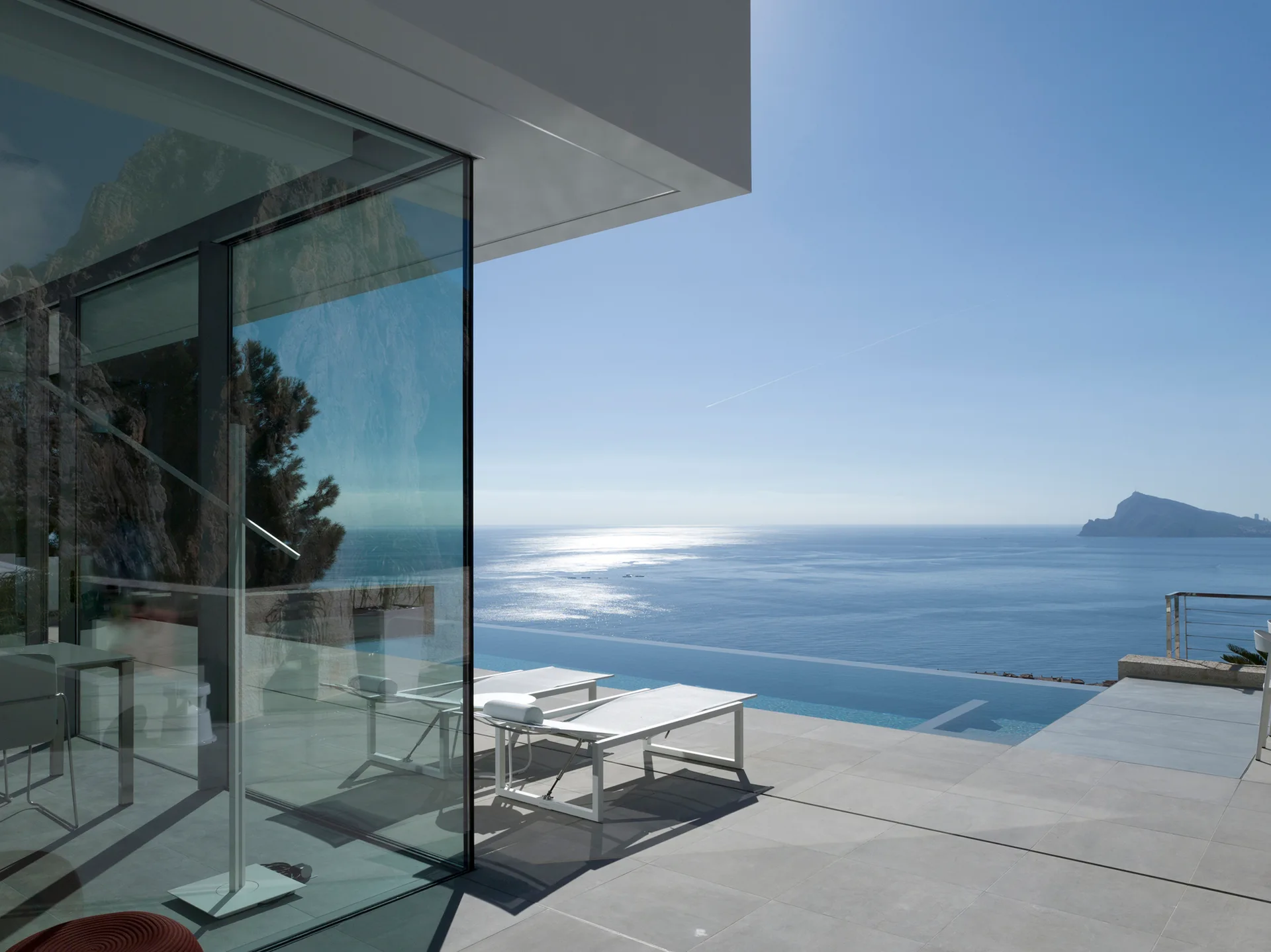 Two white sun loungers sit on a grey tiled terrace next to an infinity pool and a glass building facade overlooking the ocean.