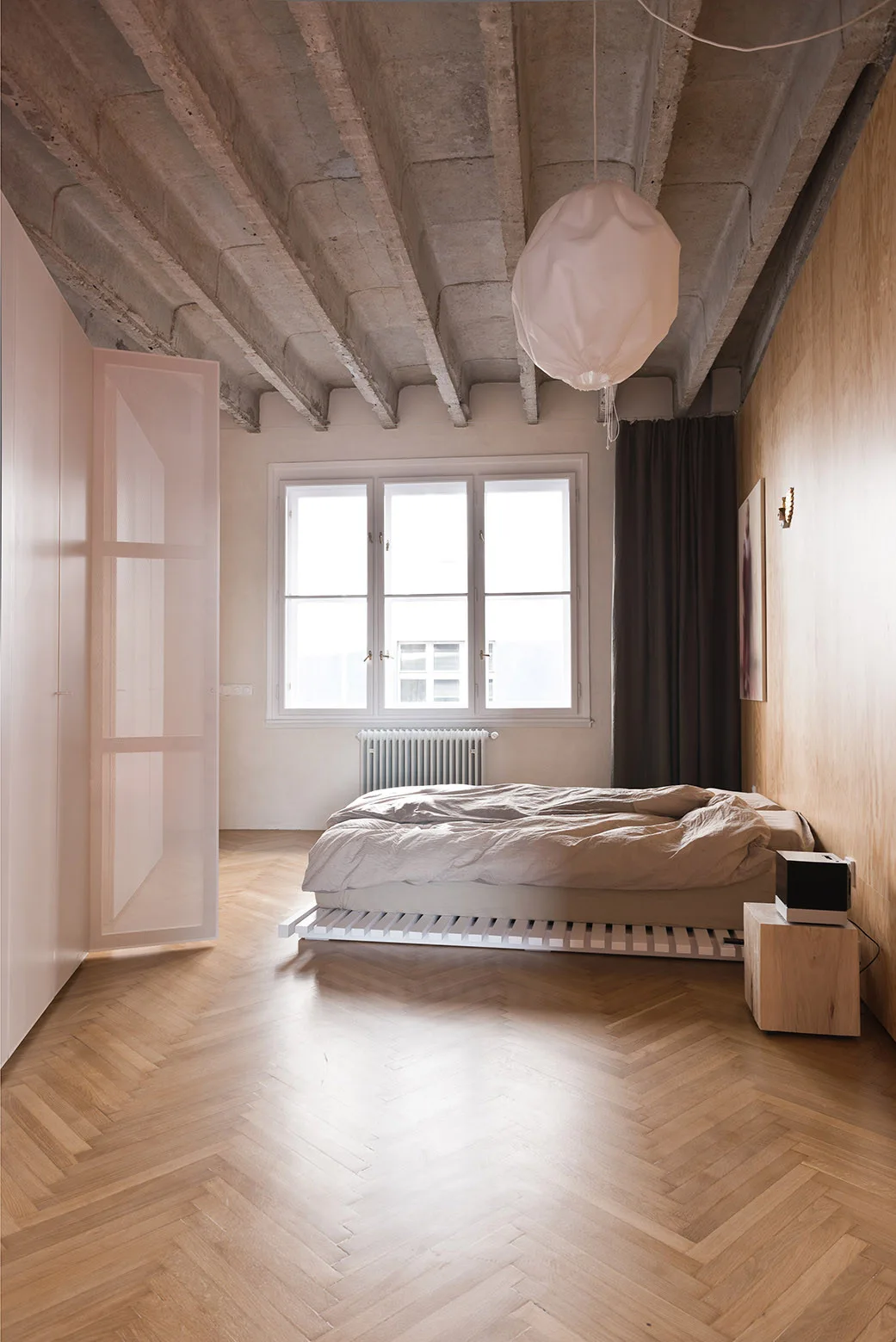 A minimalist bedroom featuring a low white slatted bed on a herringbone wood floor beneath exposed concrete ceiling beams.