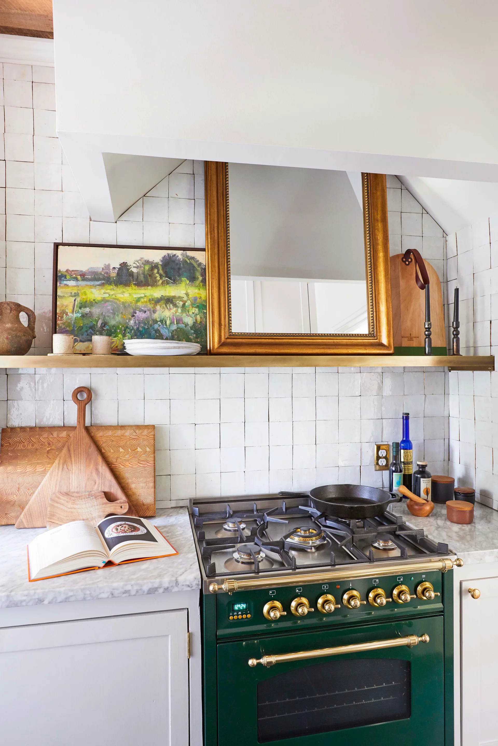 A kitchen featuring a dark green stove, a brass floating shelf with a gold-framed mirror, and a white square tile backsplash.