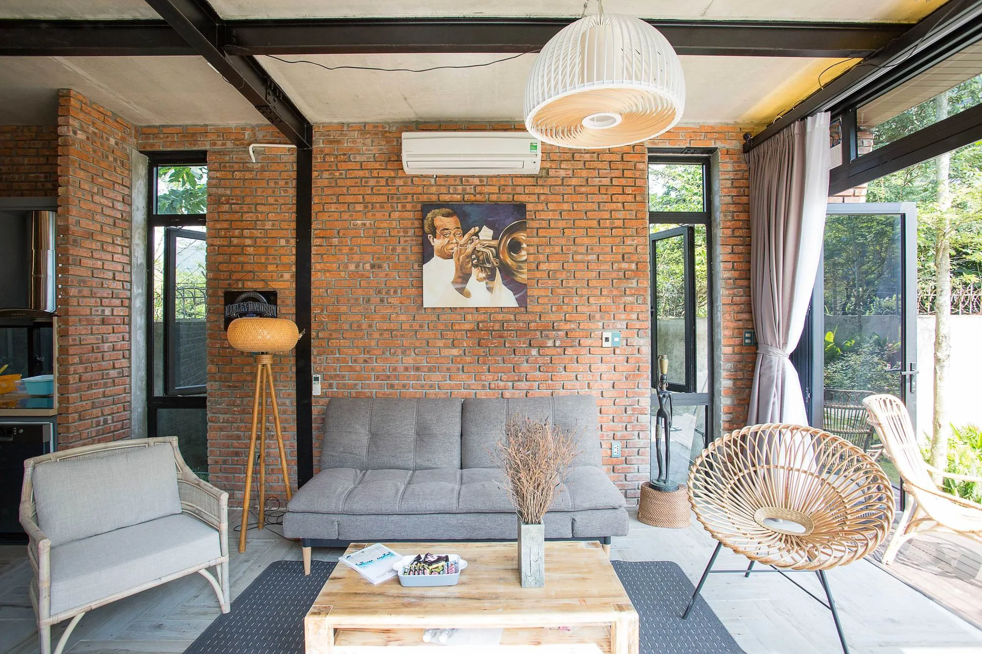 An industrial living room features a grey sofa, a wooden coffee table, and rattan chairs set against an exposed red brick wall.