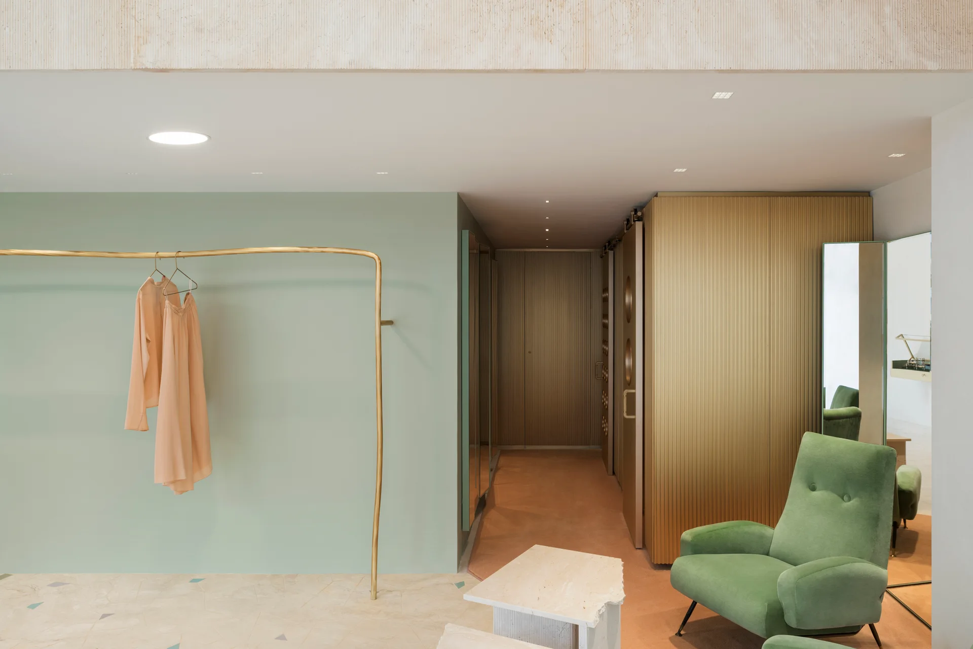 A modern dressing room features a sage green accent wall, a curved brass garment rack, a fluted gold wall partition, and a green velvet armchair.