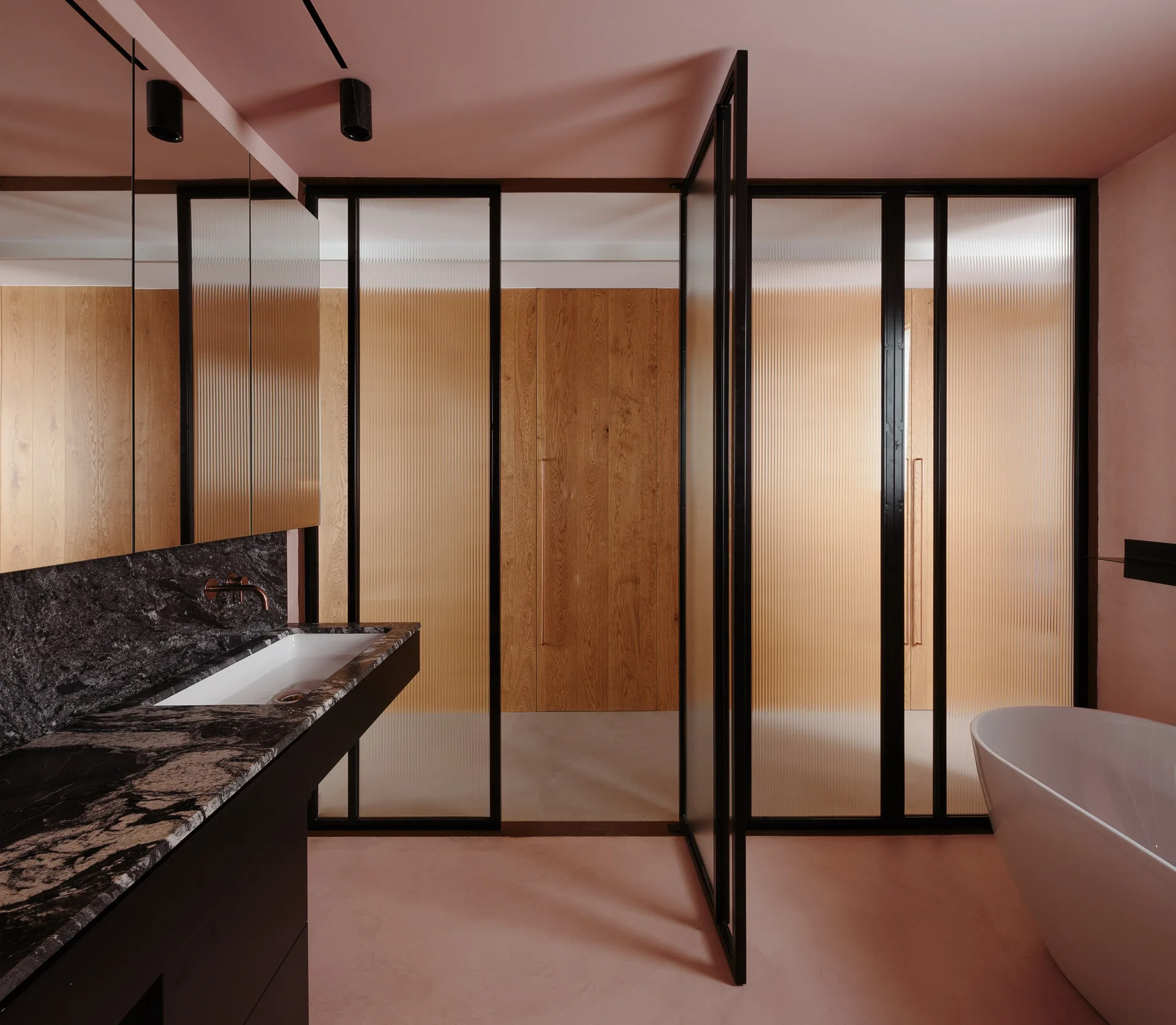 A modern bathroom with a black marble vanity, white freestanding bathtub, and pink walls separated from wood closets by black-framed fluted glass doors.