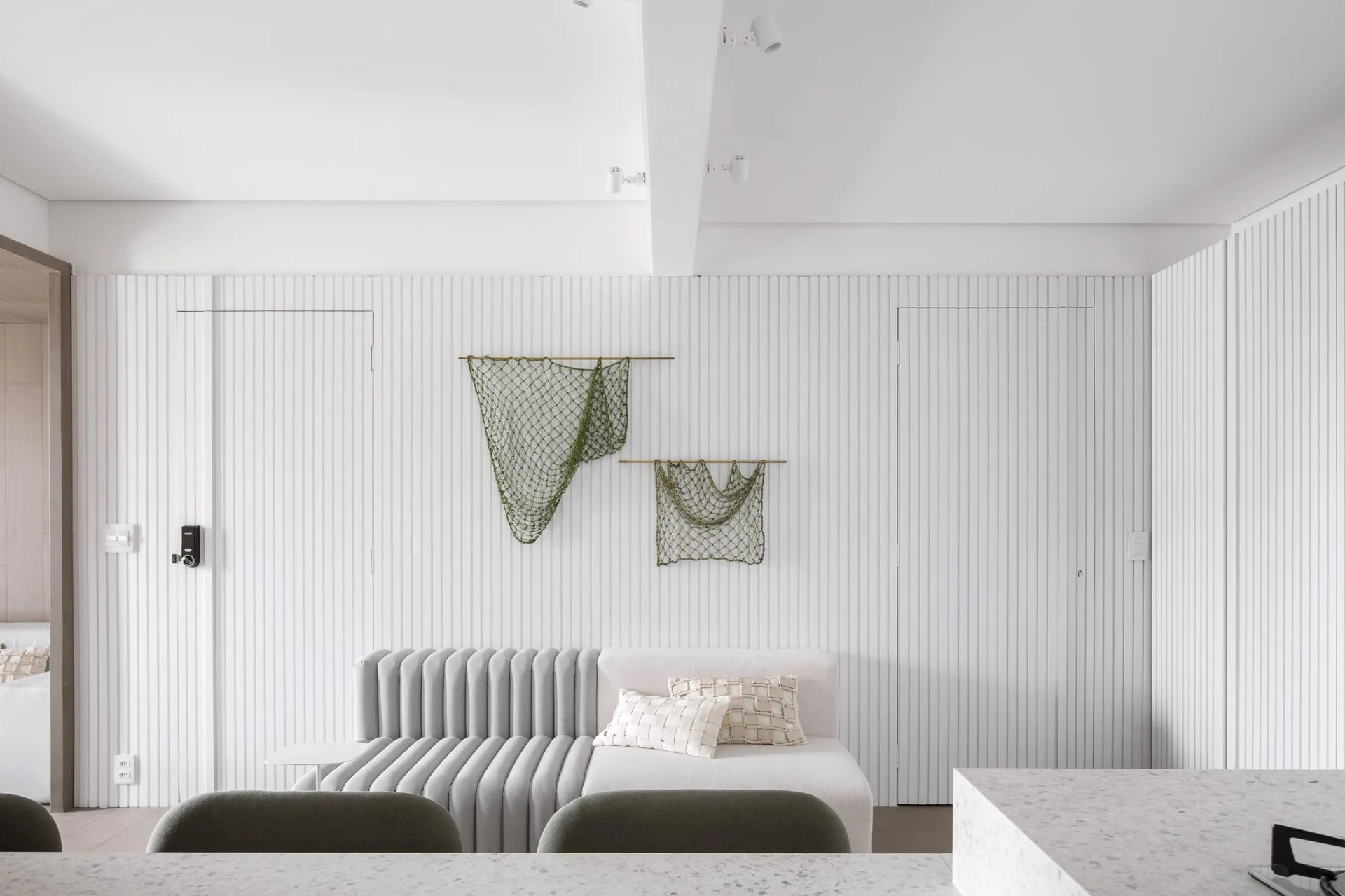 A modern white sofa sits against a vertical slatted wall featuring two hidden doors and green netting wall art.