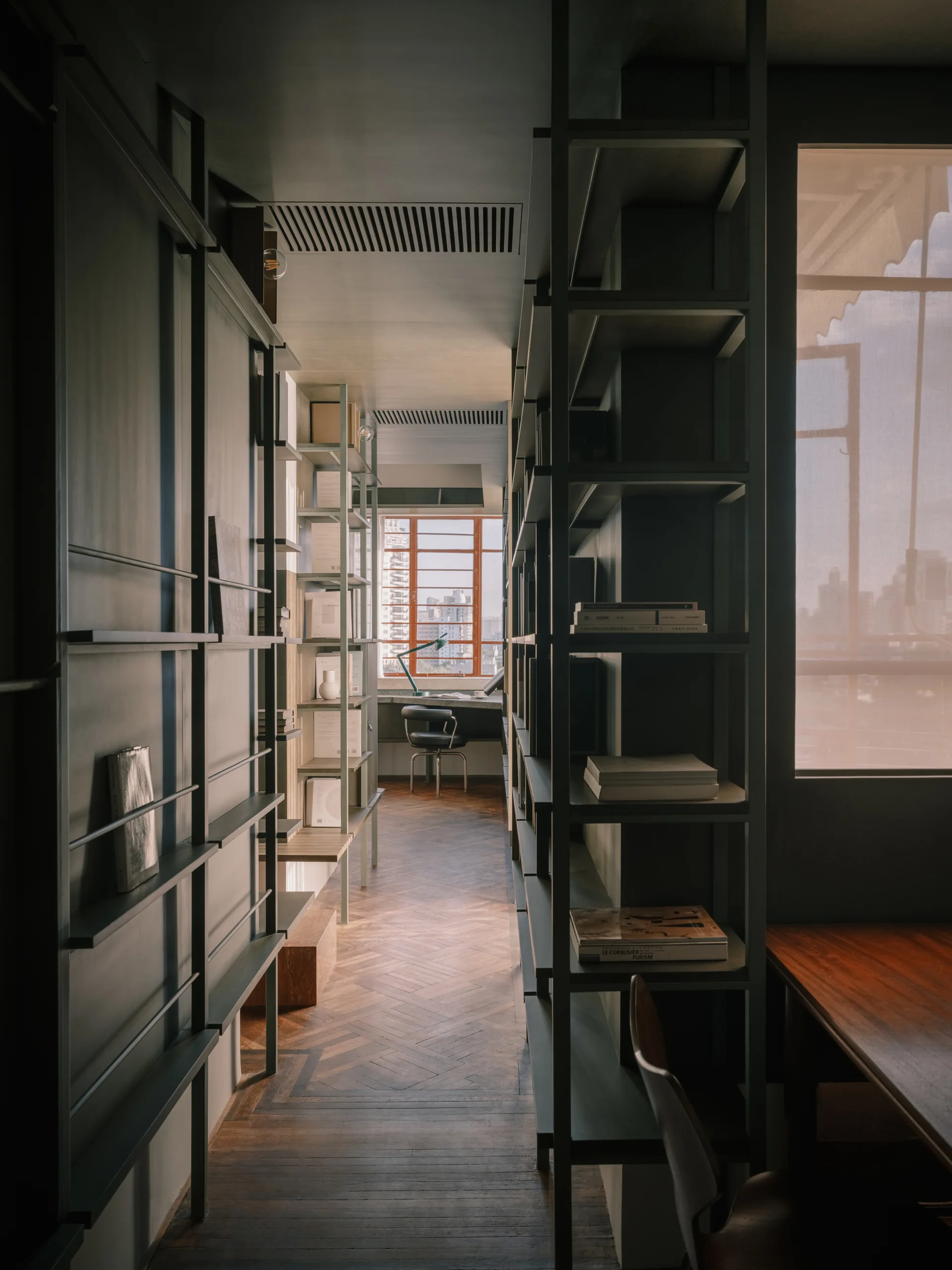 Tall dark gray metal bookshelves line a narrow corridor with herringbone wood floors leading to a bright desk area by a window.