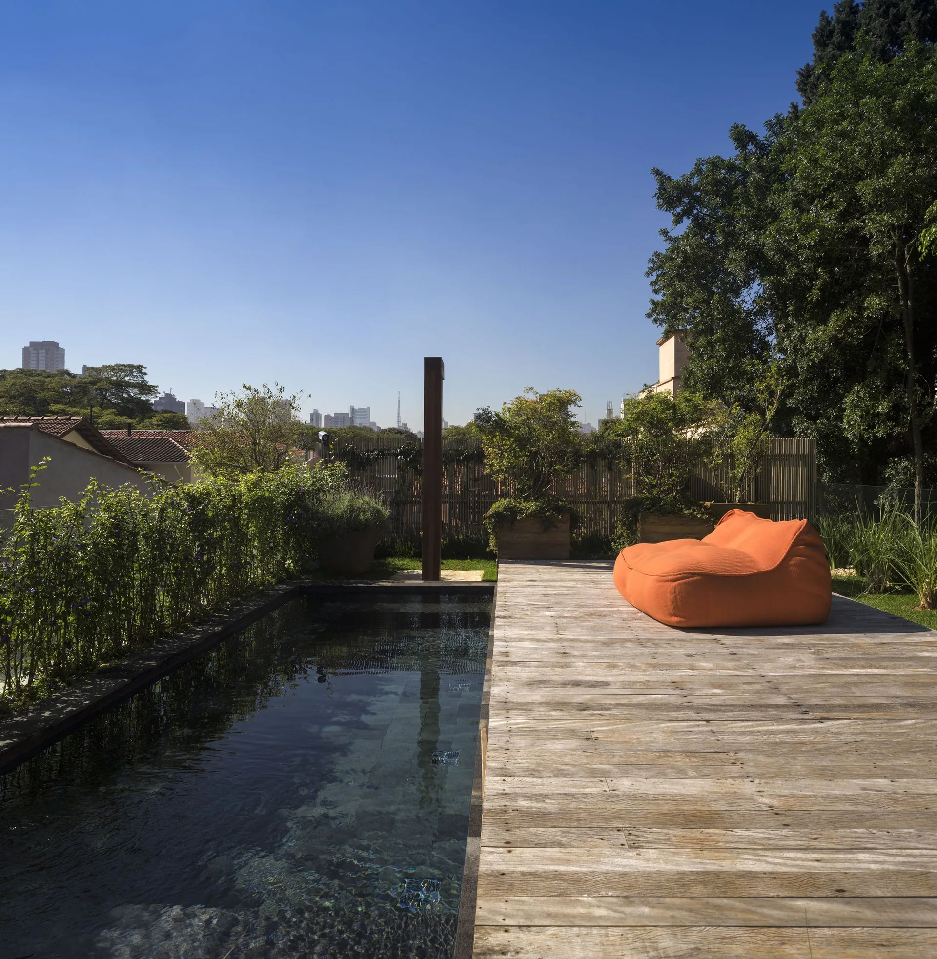 A rooftop terrace features a rectangular swimming pool next to weathered wood decking, a bright orange beanbag lounge chair, and green hedges.