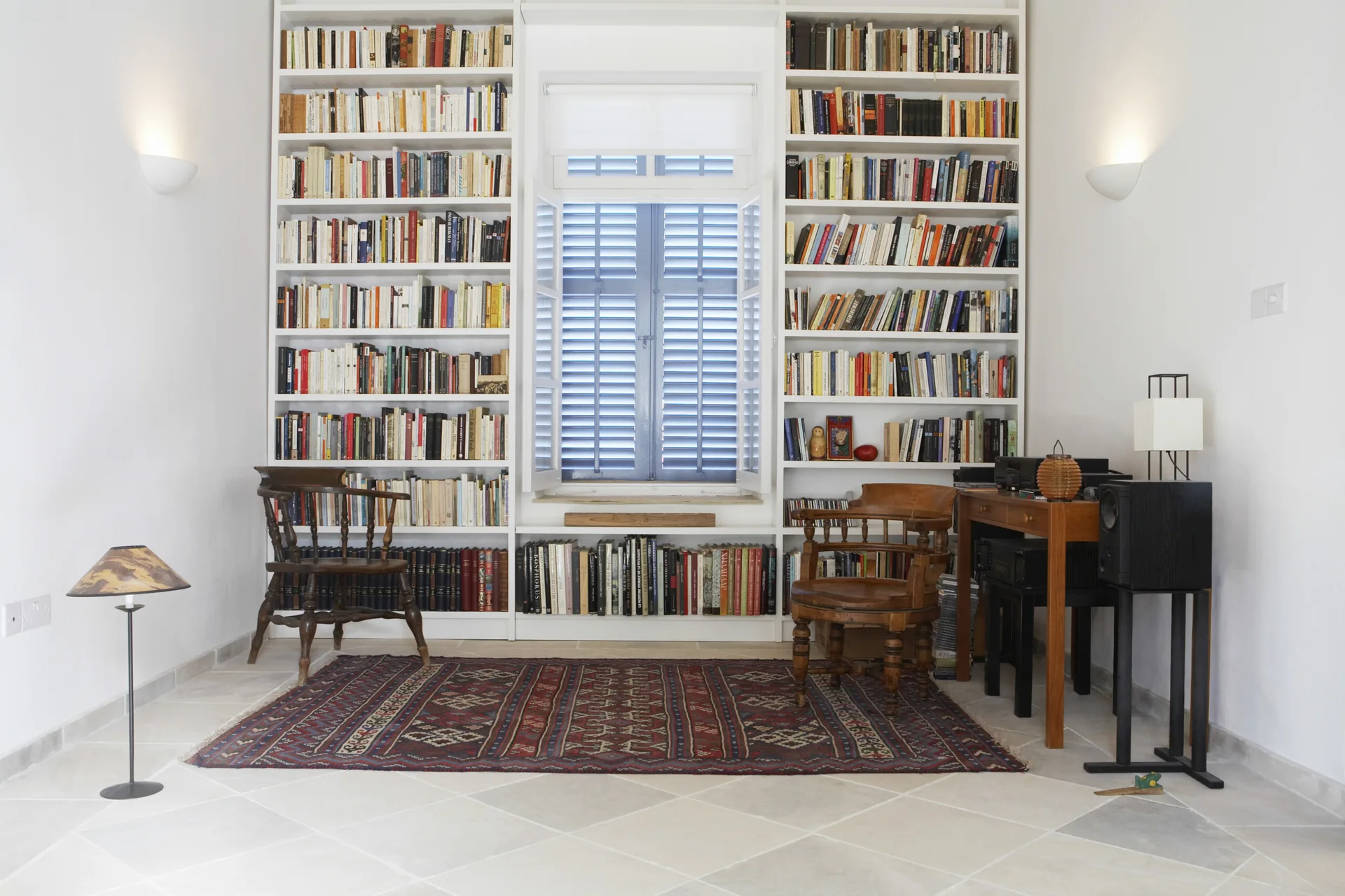 A home library features built-in white bookshelves flanking a window with blue shutters, two wooden spindle chairs, and a patterned rug on a stone tile floor.