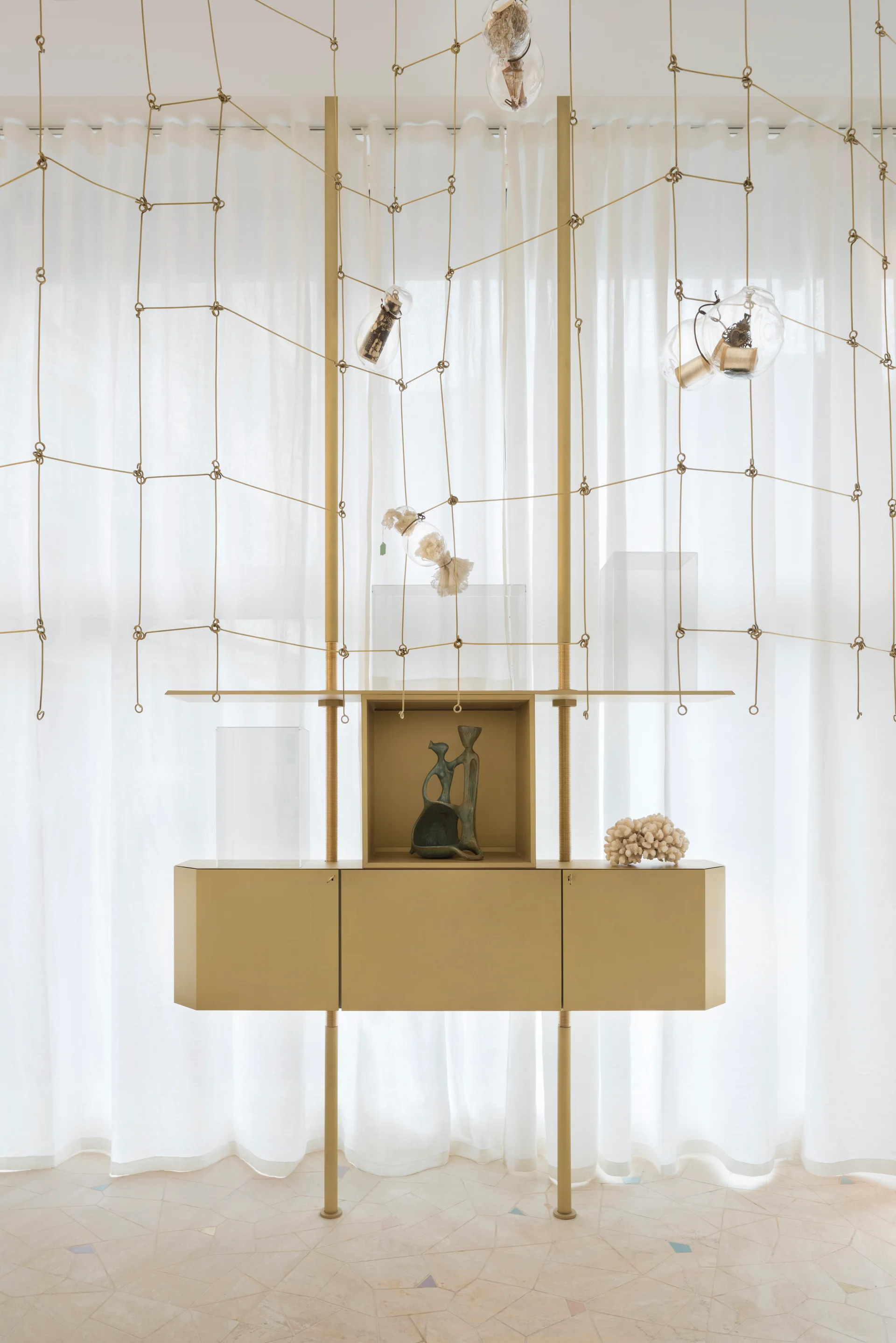 A gold metal display cabinet stands against sheer white curtains, featuring a suspended rope net adorned with hanging glass ornaments.