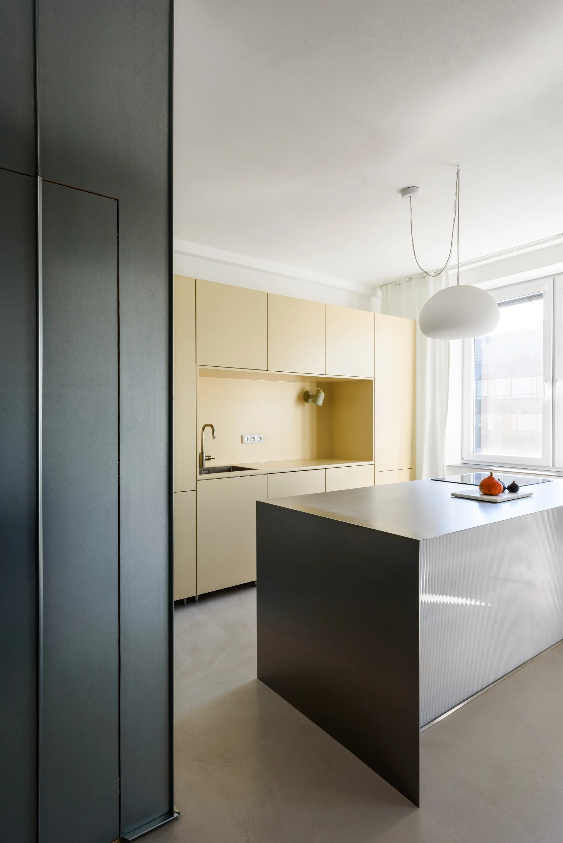 A minimalist kitchen features matte pale yellow built-in cabinetry, a dark gray metal kitchen island with a fluted side panel, and a white pendant light.