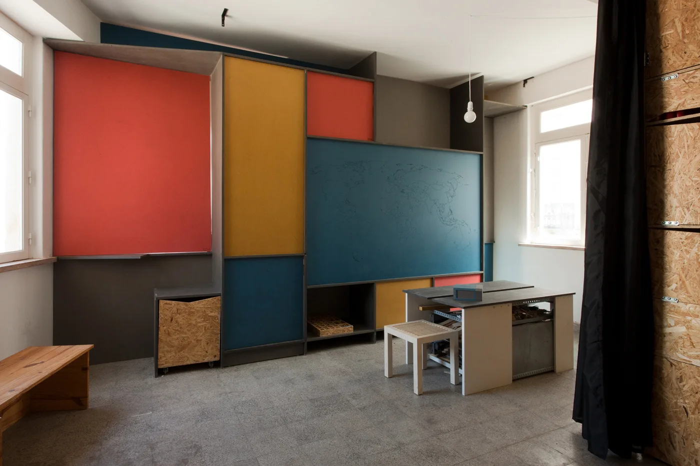 A modern creative workspace features a large multicolored modular wall cabinet, a central gray desk with a wooden stool, and a speckled gray floor.