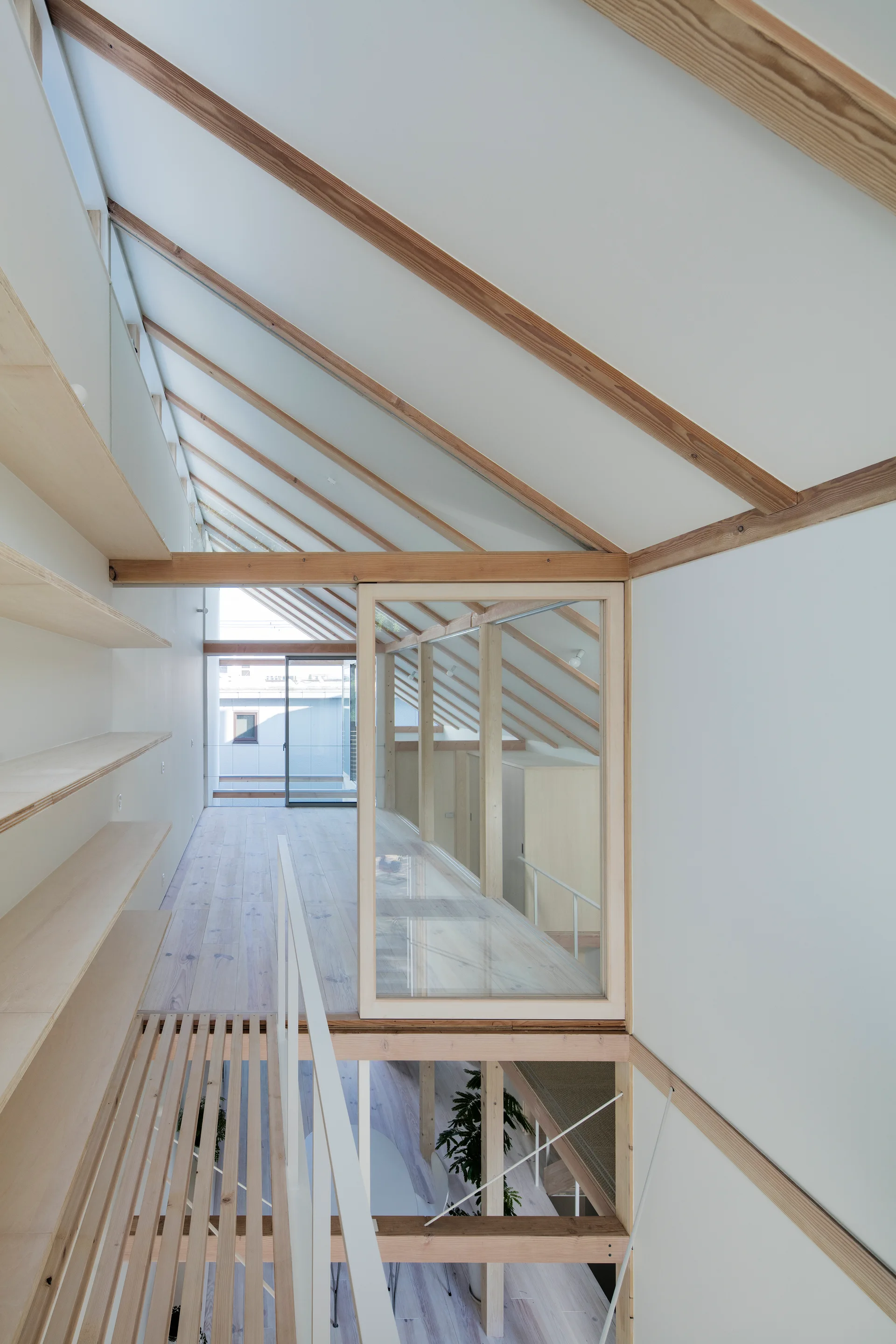 A minimalist loft landing features a sloped ceiling with exposed wood beams, built-in plywood shelving, and a large glass partition.