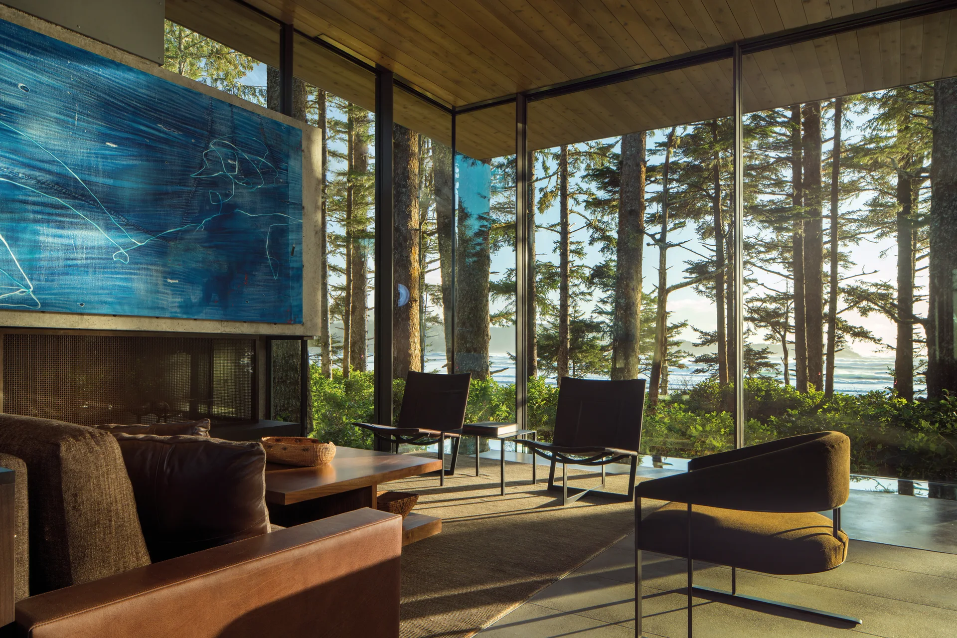 Modern living room with floor-to-ceiling glass walls showing a pine forest, furnished with low-profile leather seating and a wood plank ceiling.