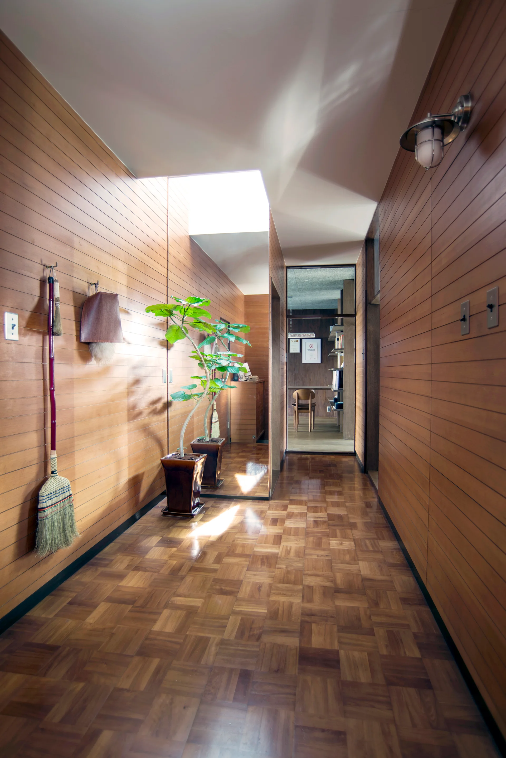 A narrow hallway features warm horizontal wood-paneled walls, parquet wood flooring, a ceiling skylight, and a potted plant next to a large mirror.