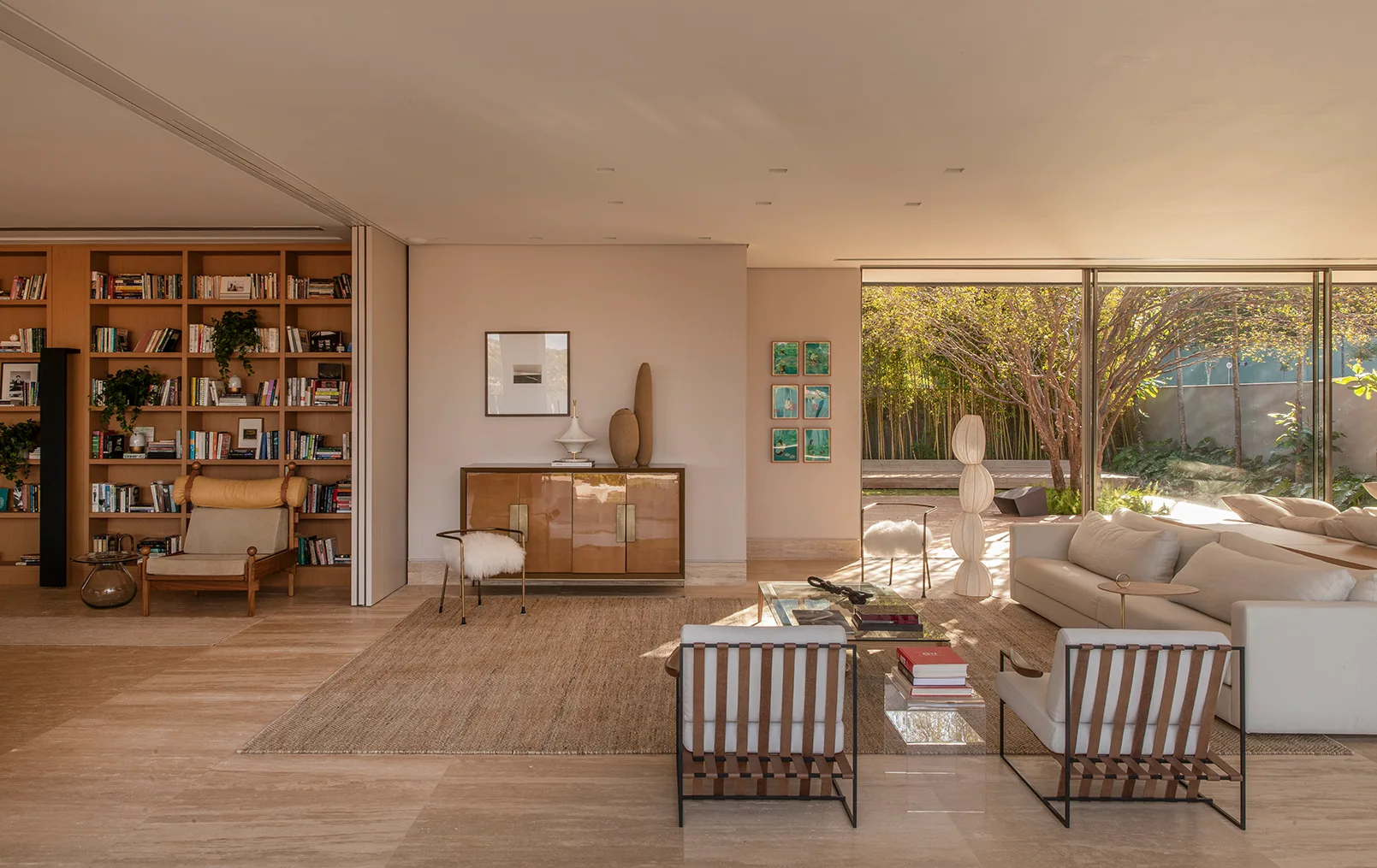 A warm neutral living room features a built-in wooden bookcase, neutral seating, a woven rug, and a large sliding glass door opening to a garden.