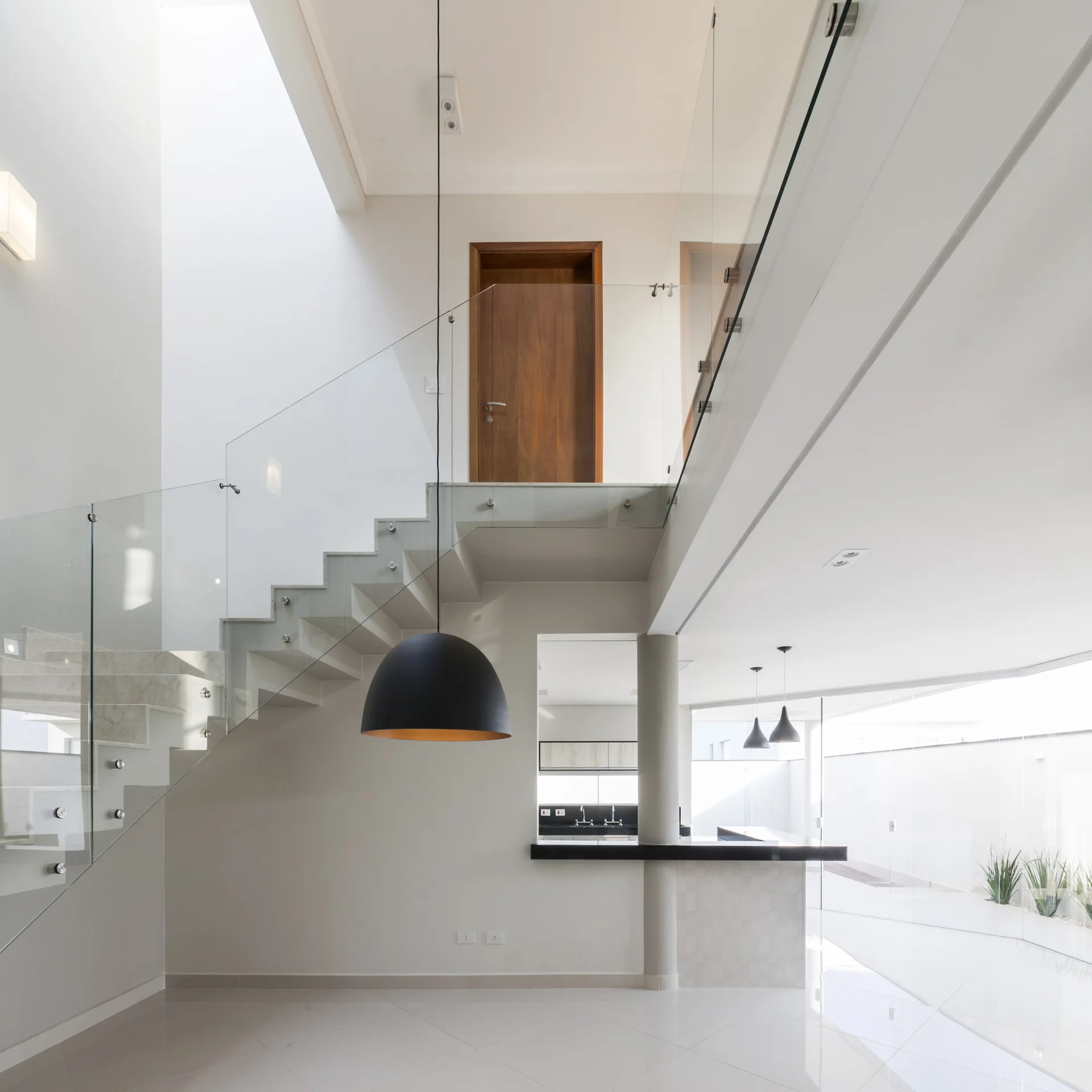 A modern foyer features a floating concrete staircase with a glass railing, white walls, polished tiled flooring, and a large black pendant light.