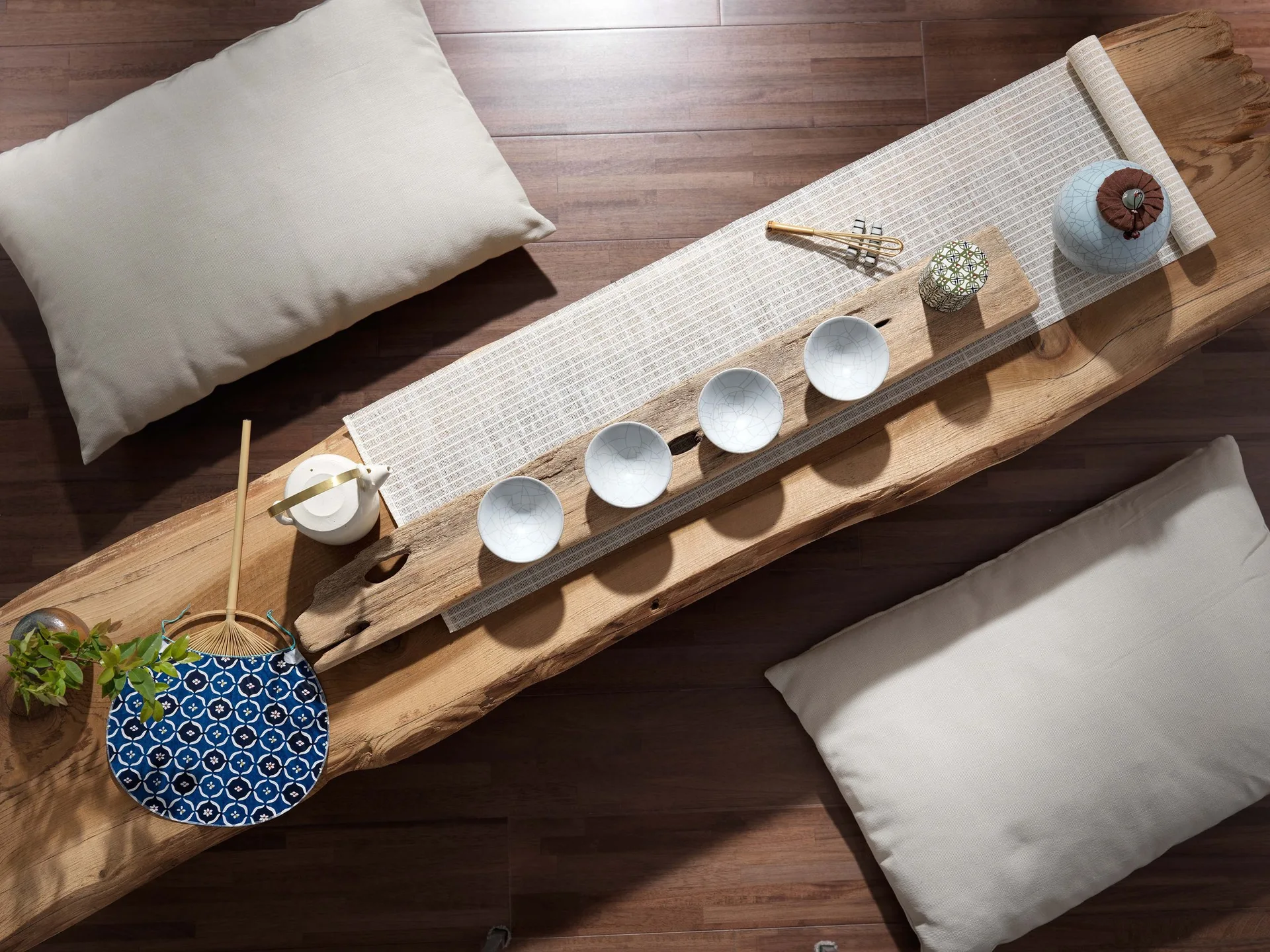 Overhead view of a tea ceremony arrangement set on a live-edge wooden bench with four ceramic cups, a teapot, and floor cushions.