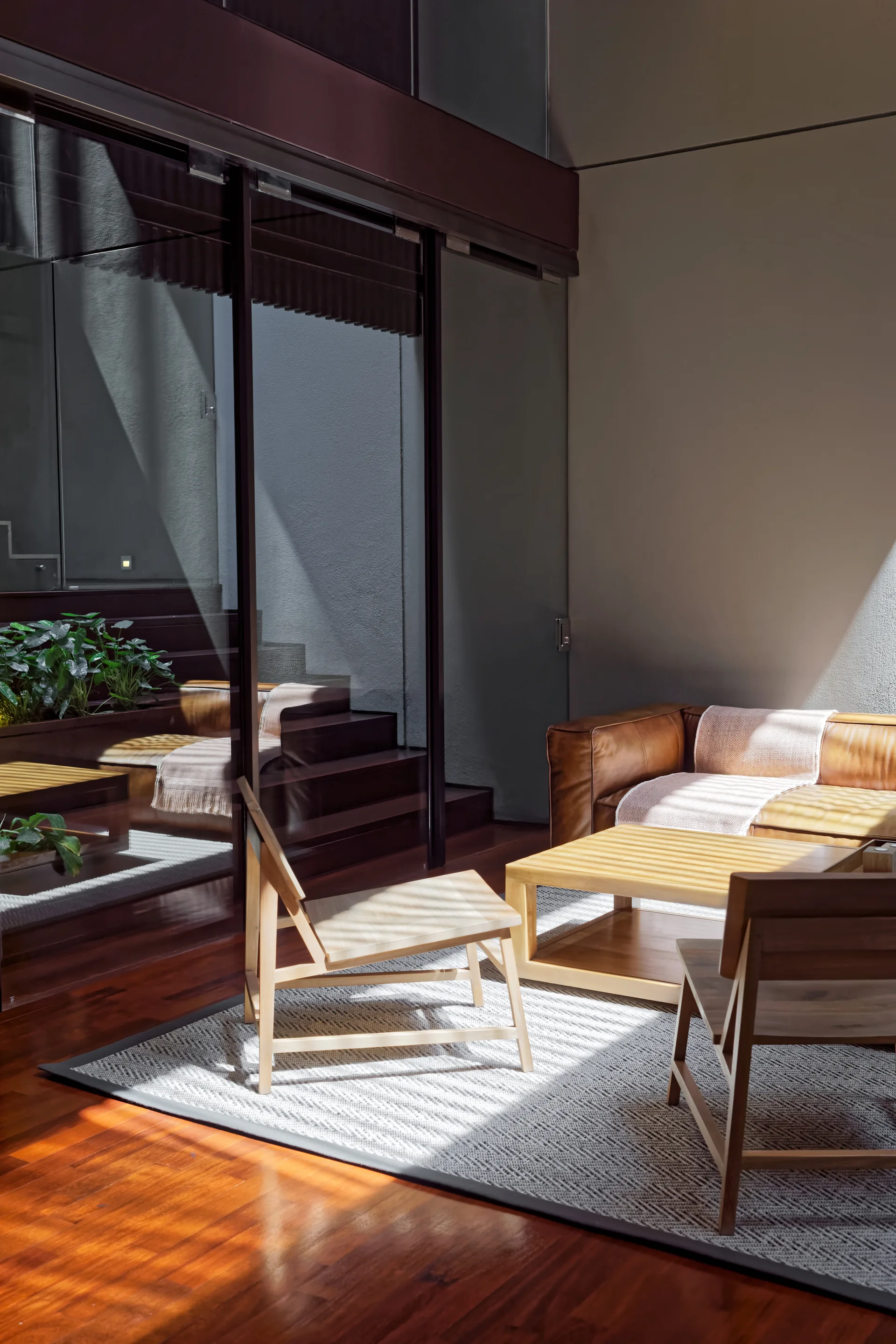 A sunlit living area features a tan leather sofa, low wooden chairs, a geometric patterned rug, and a dark-framed glass partition revealing steps.