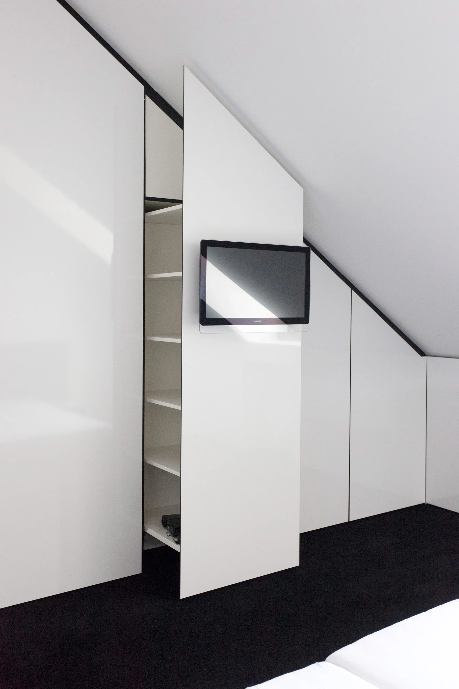 A glossy white custom wardrobe built into a sloped attic ceiling features a television mounted on one of the door panels.