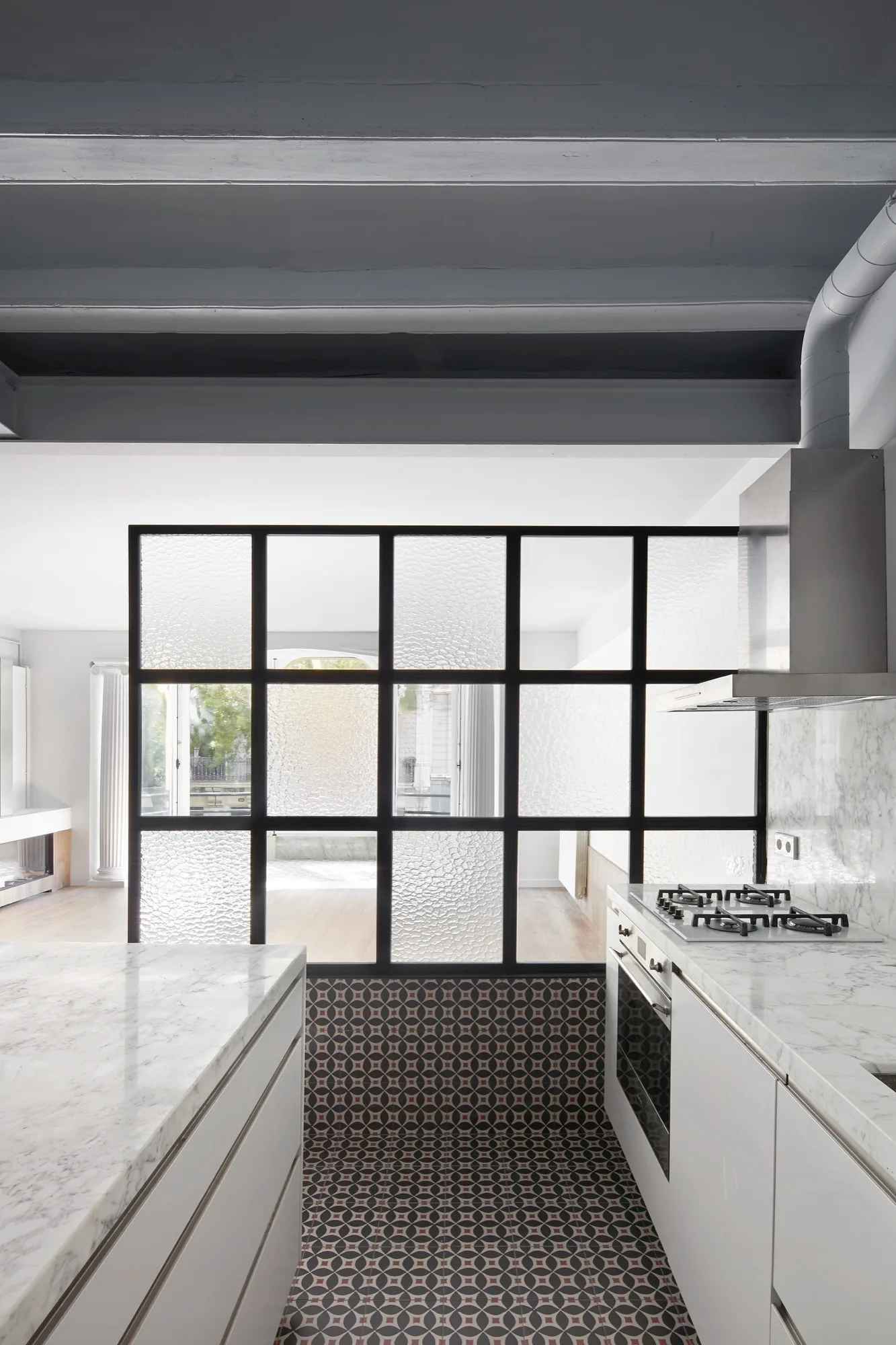 A modern kitchen features white marble countertops, a black metal-framed textured glass partition, and geometric patterned floor tiles.