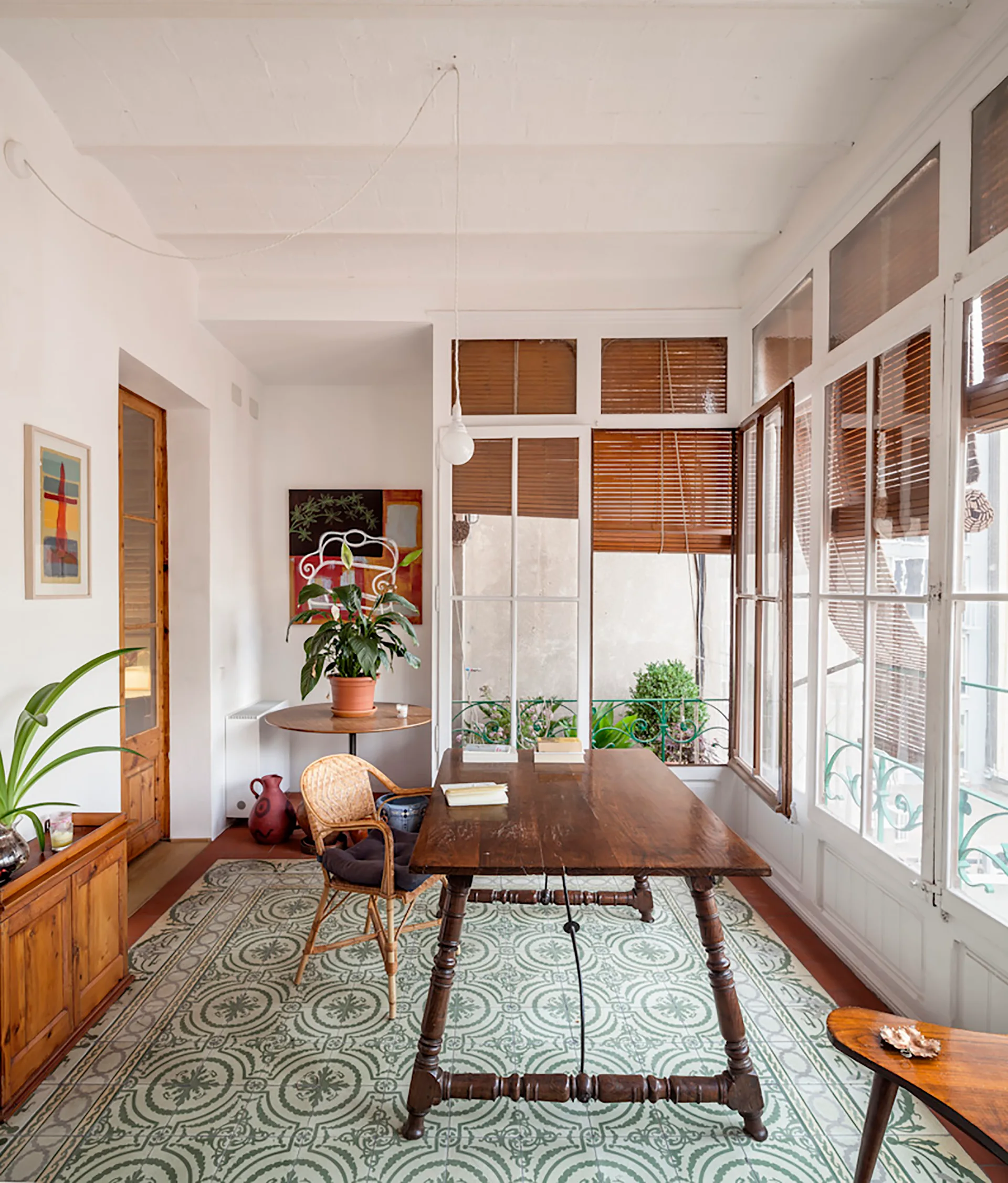 A bright sunroom office features a rustic wooden table and wicker chair atop green and white patterned tile flooring under large windows with wooden blinds.