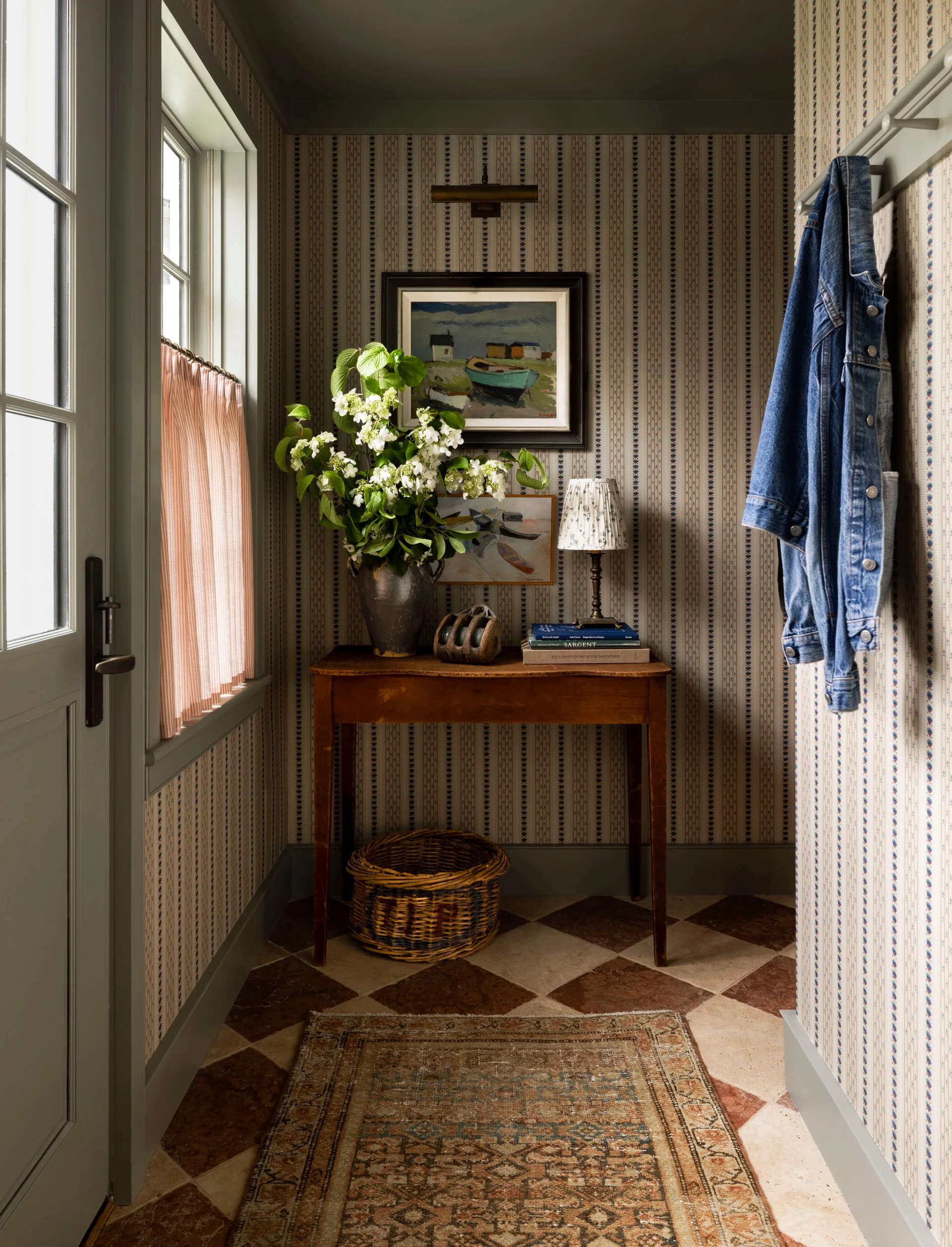 A cottage entryway features vertical striped wallpaper, a checkered stone tile floor, and an antique wooden console table decorated with white flowers.