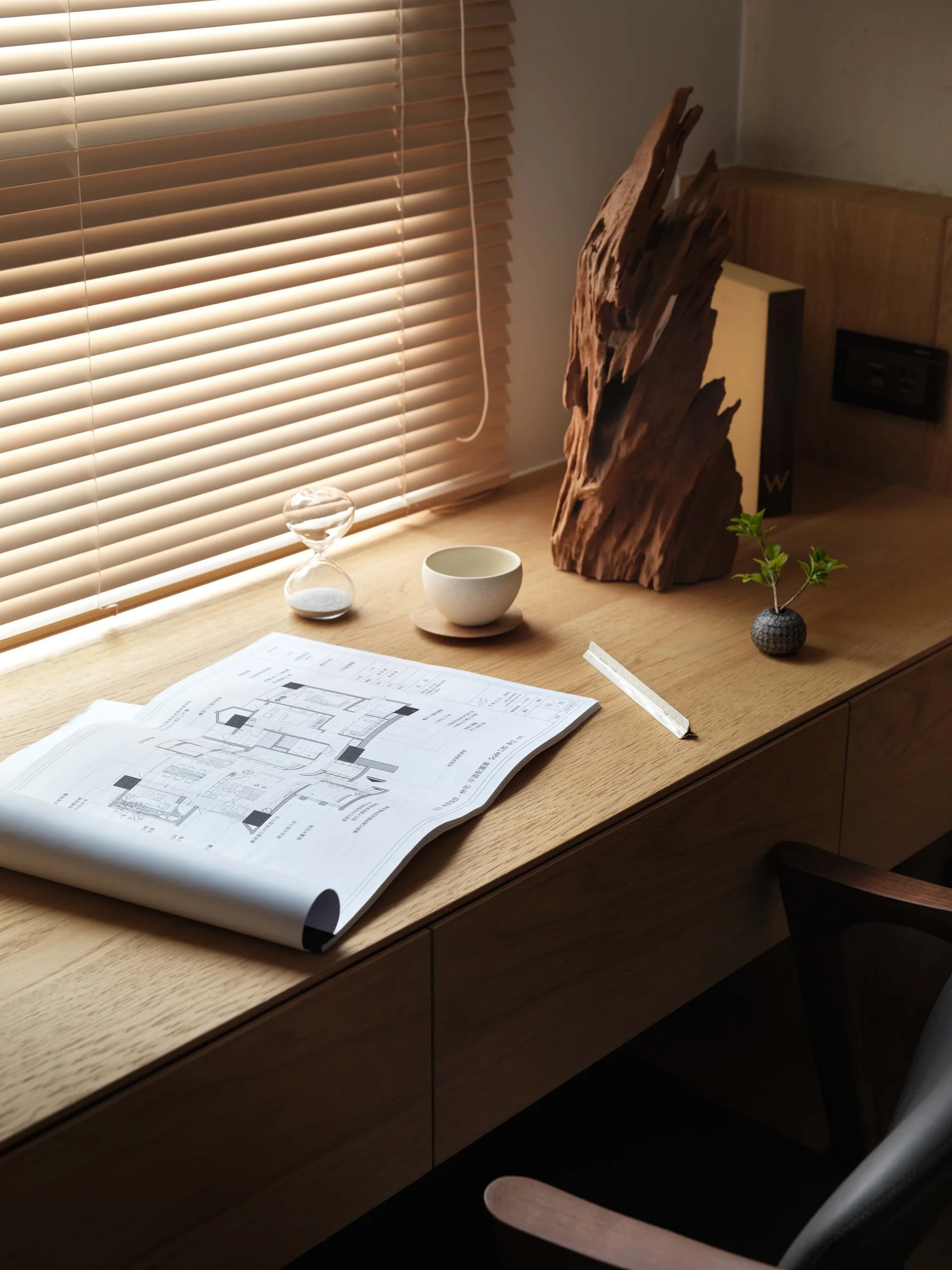 An architectural drawing lies on a light wood desk next to a rustic wood sculpture under soft light filtering through window blinds.