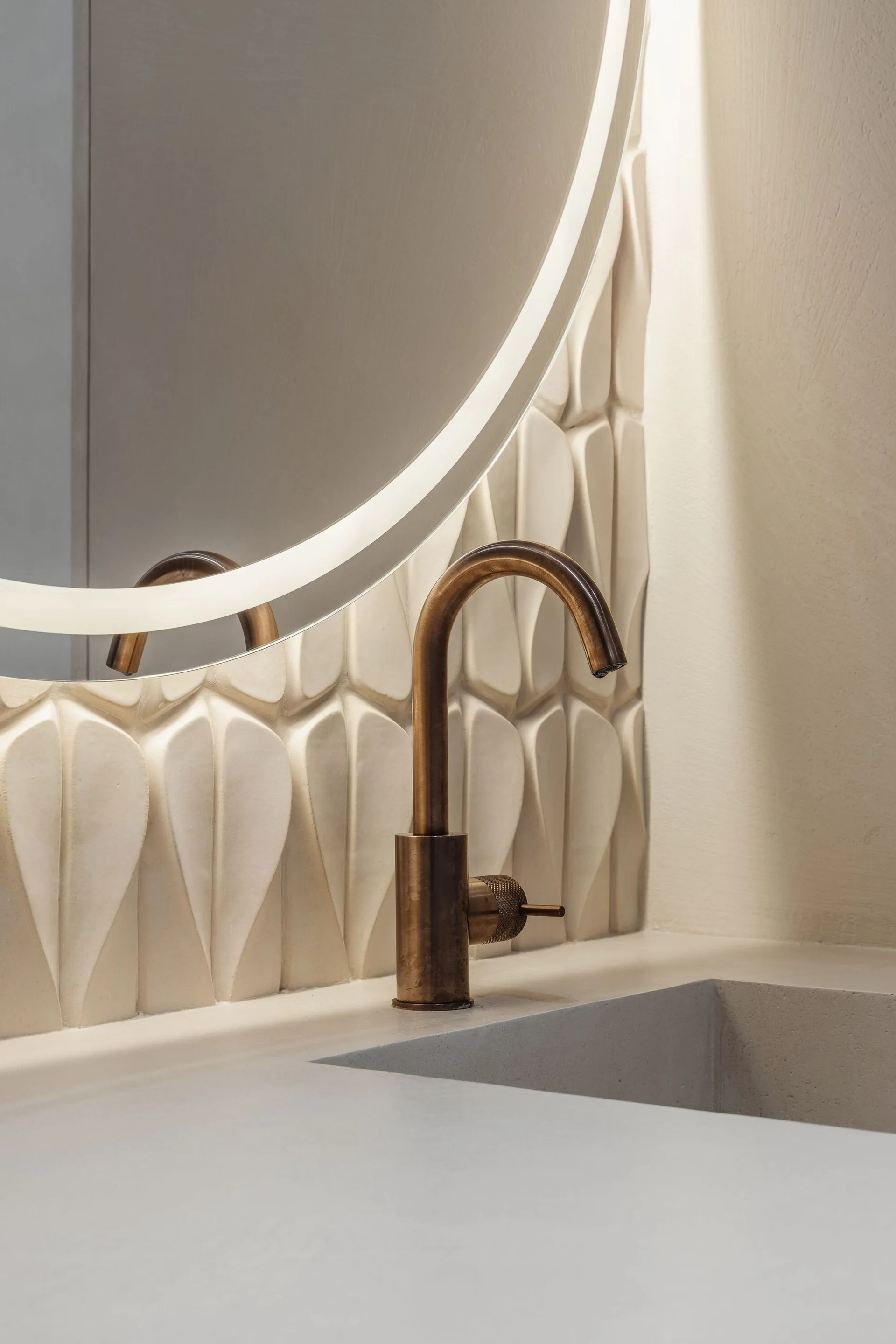 A close-up of a bathroom sink with a bronze faucet in front of a dimensional leaf-patterned tile wall and a circular backlit mirror.