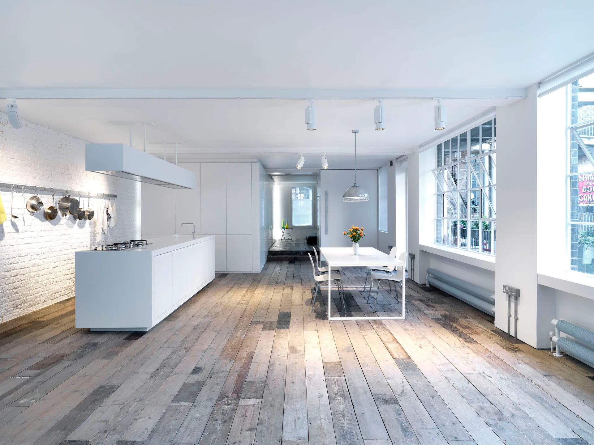 A minimalist kitchen featuring a white island, white-painted brick walls, a simple dining set, and worn wide-plank wood flooring.