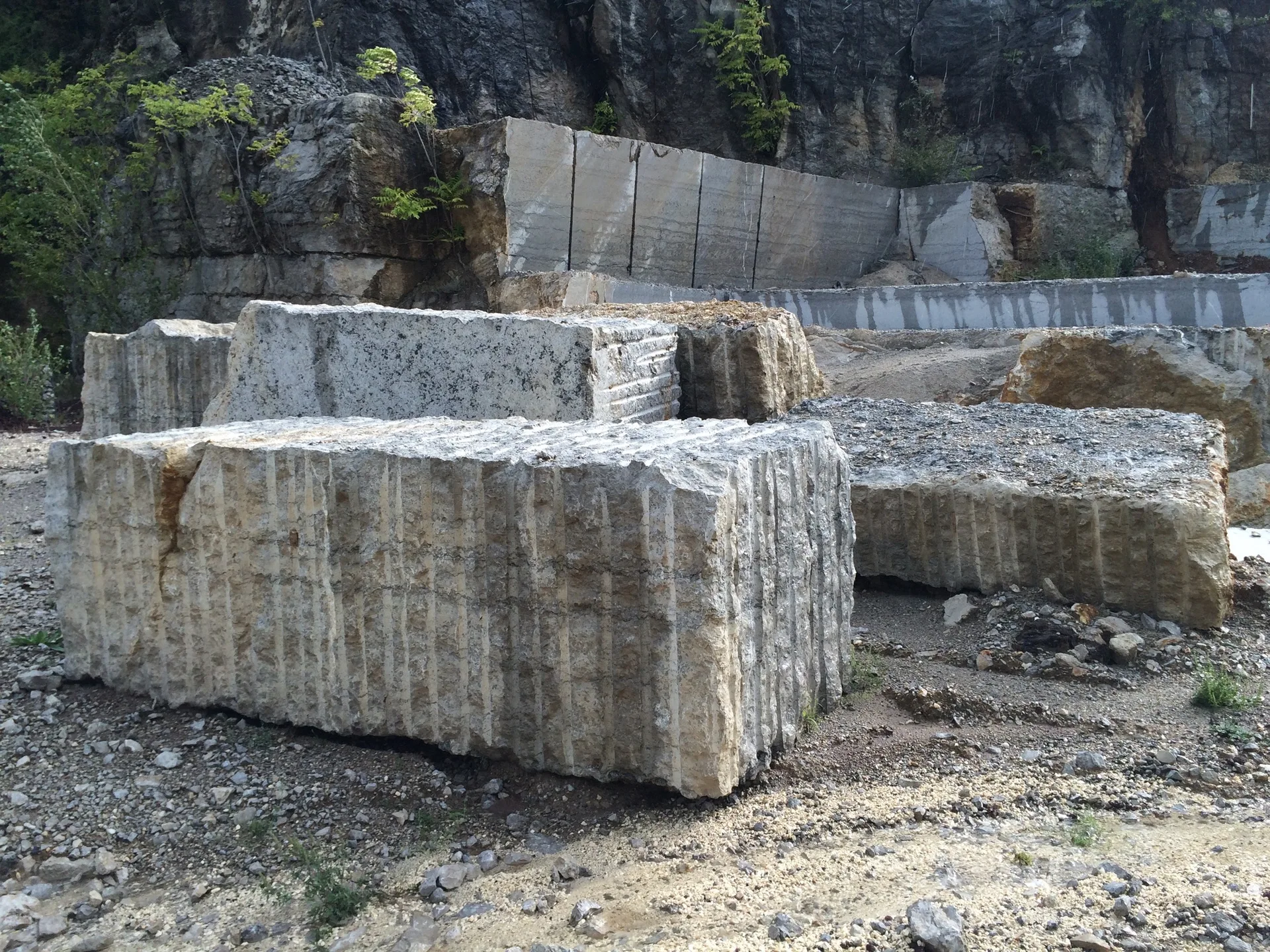 Large rectangular grey stone blocks with vertical grooves sit on a gravel ground inside an outdoor quarry with a rock cliff background.
