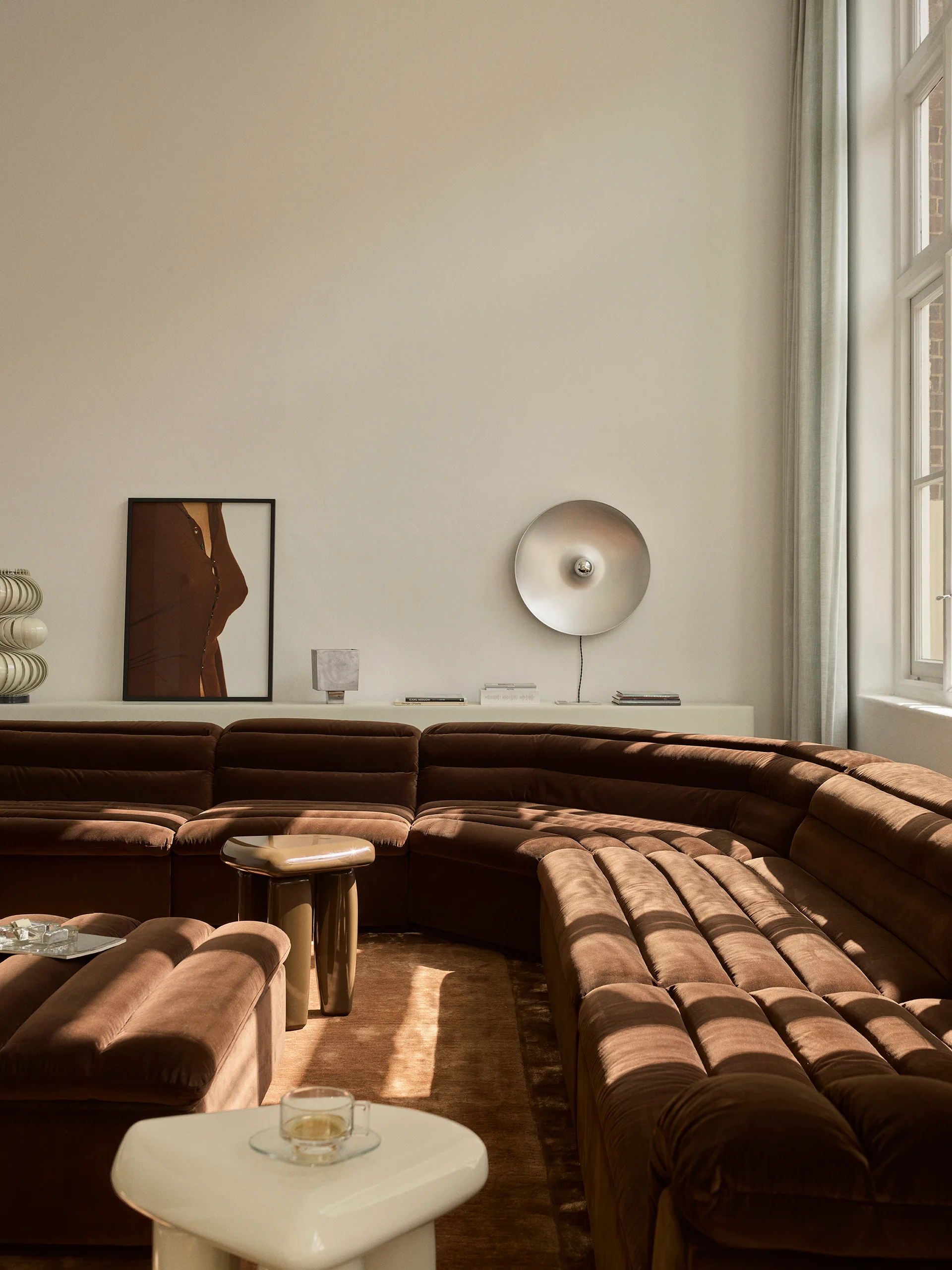 A minimalist living room featuring a chocolate brown velvet channel-tufted sectional sofa, sculptural side tables, and a low display ledge on the wall.
