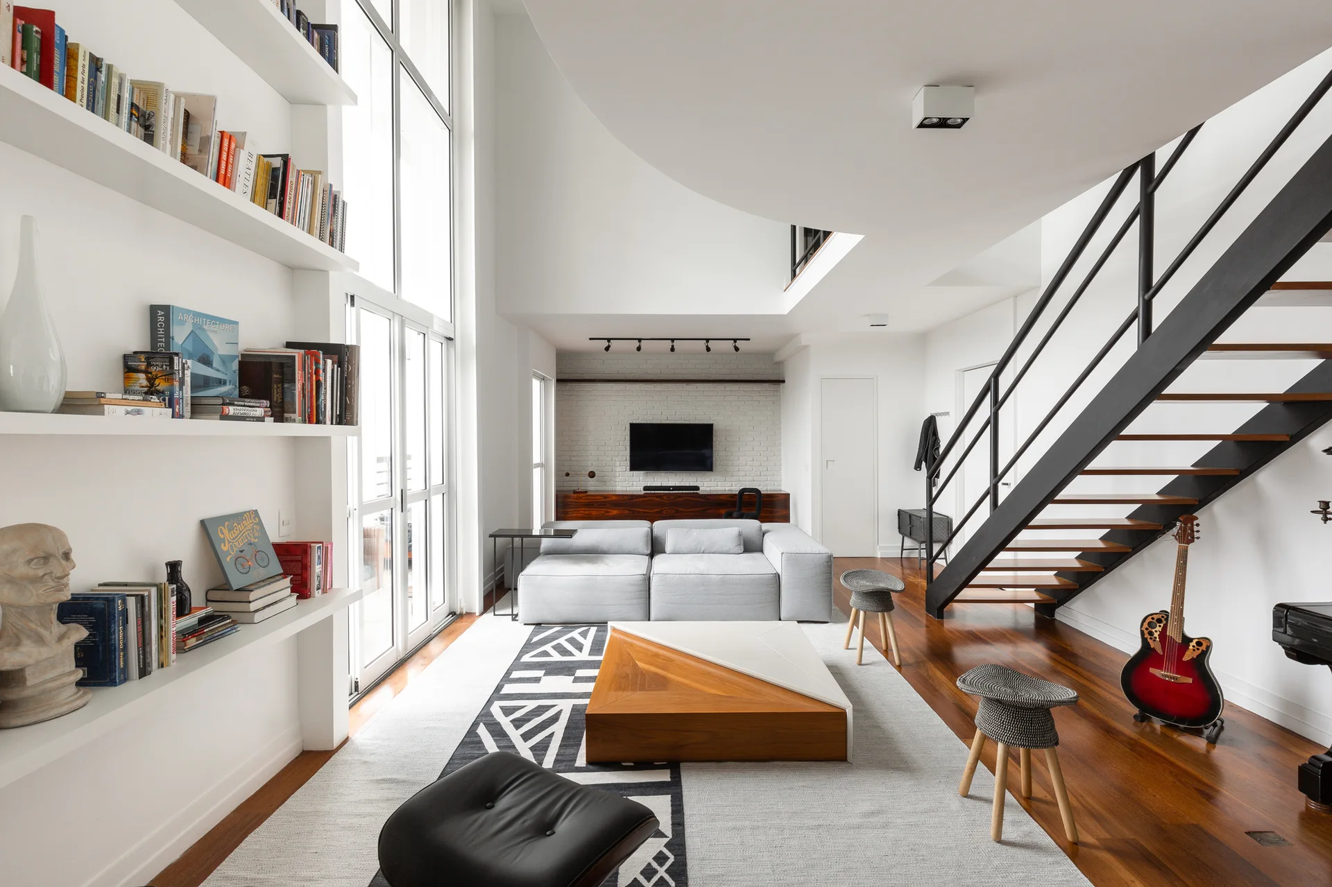 A double-height living room featuring a light gray modular sofa, a geometric wood coffee table, built-in bookshelves, and an open metal staircase.