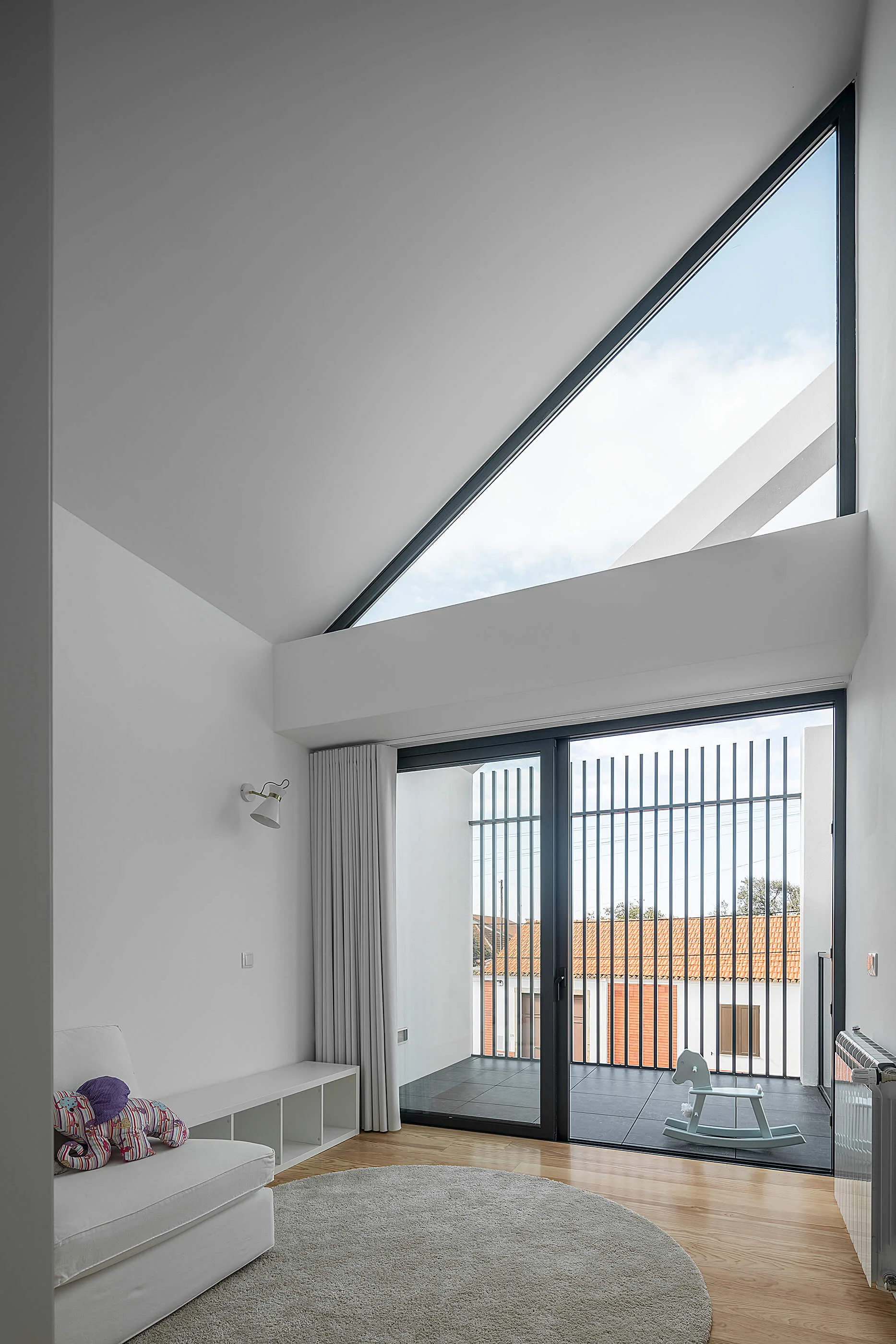 A bright minimalist room featuring a triangular window, a sliding glass door leading to a balcony, a white sofa, and light wood flooring.