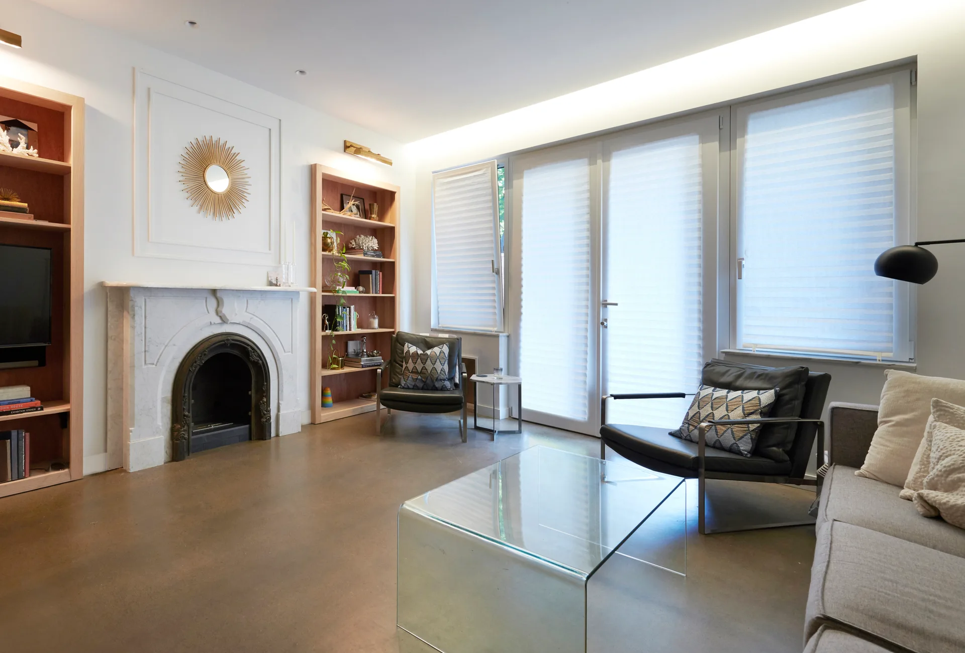 A contemporary living room featuring a white marble fireplace, light wood built-in bookcases, black leather armchairs, and a glass coffee table.