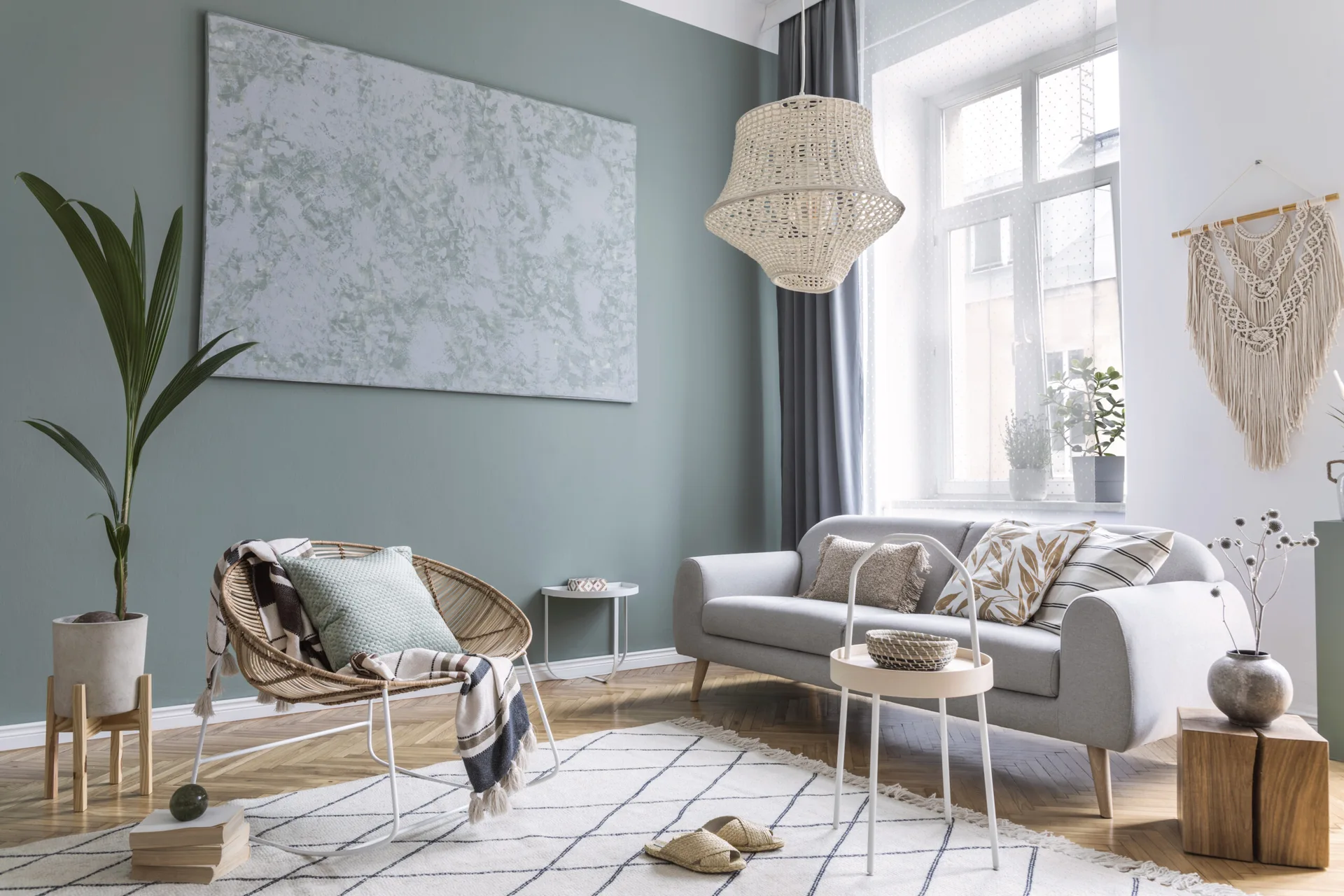 A Scandi-style living room features a sage green accent wall with large art, a gray sofa, a rattan rocking chair, and a woven pendant light.