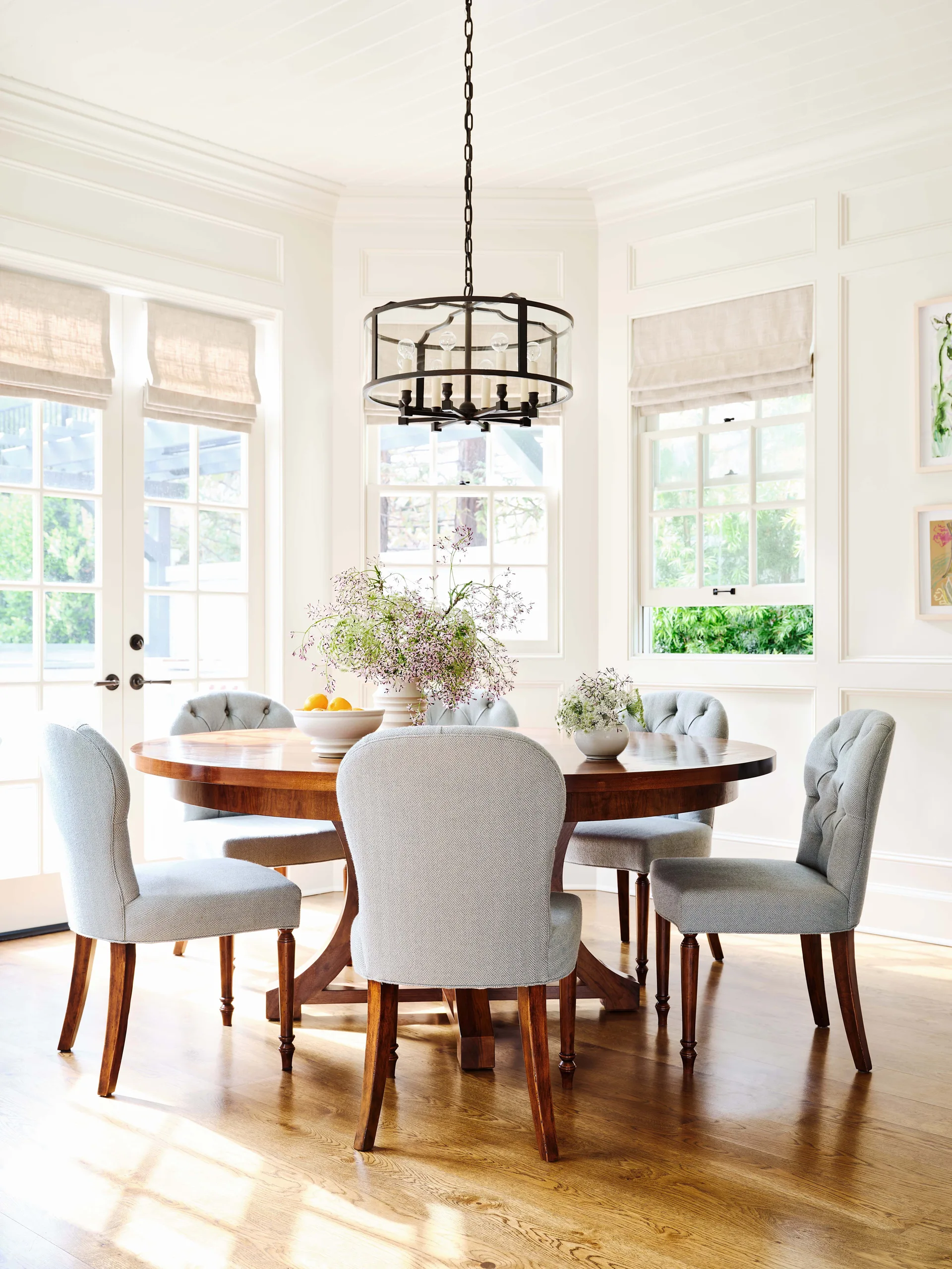 A bright dining room features a round wooden pedestal table surrounded by light blue upholstered chairs beneath a dark circular metal chandelier.