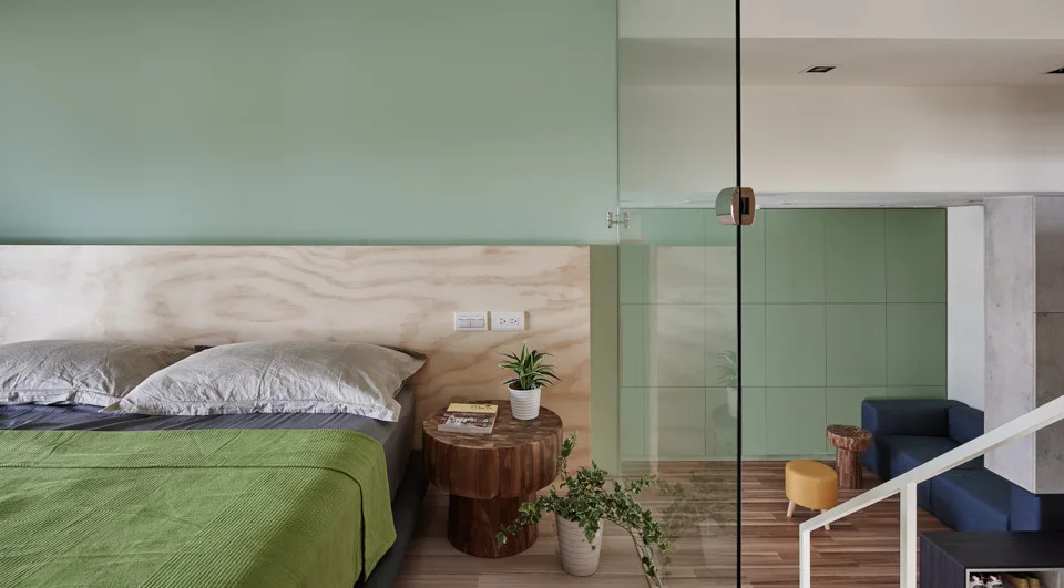 A modern sage green bedroom features a natural wood headboard and a glass wall divider overlooking an adjacent lounge with a blue sofa.
